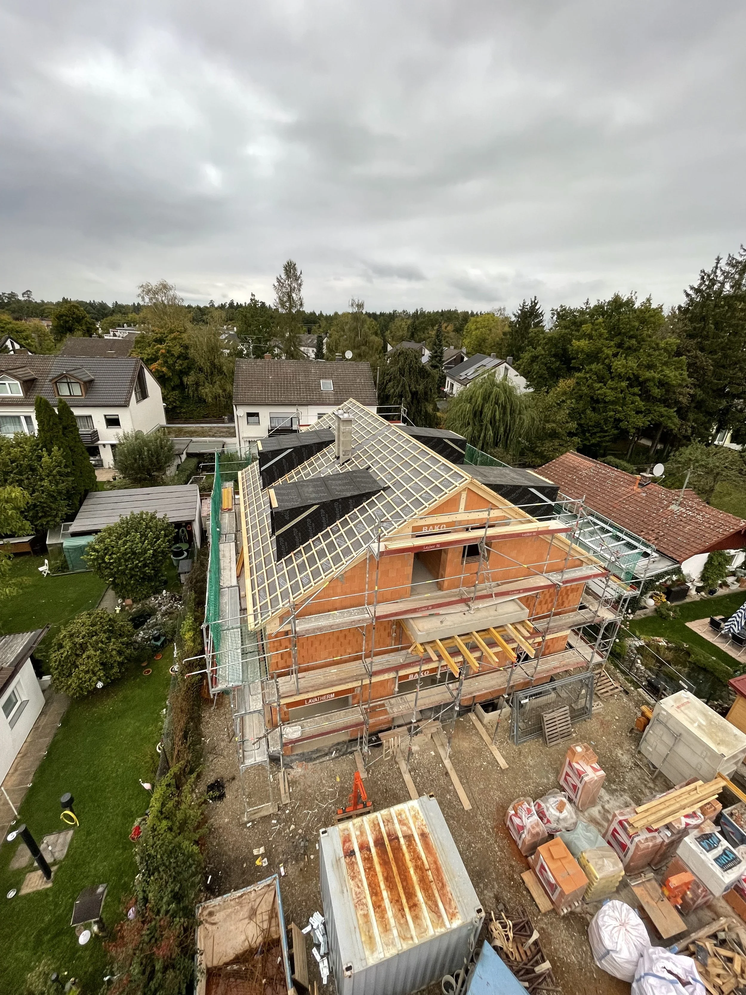 A house under construction with scaffolding and building materials on a cloudy day, surrounded by trees and neighboring houses