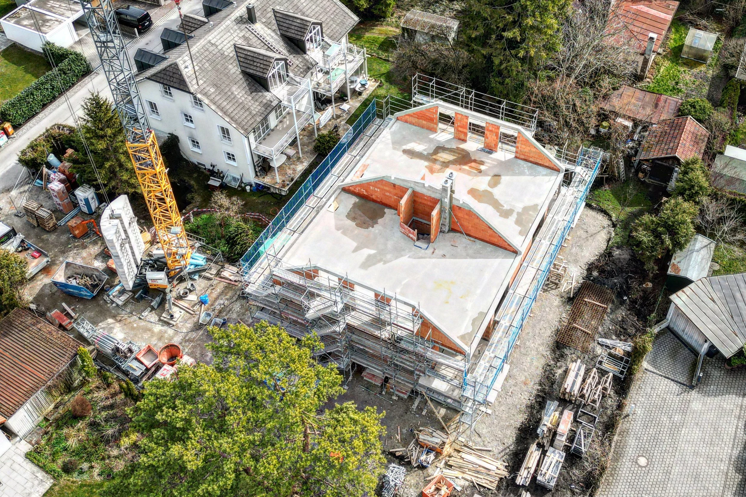 Aerial view of a construction site with a partially built multi-story building, scaffolding, construction materials, a crane, and surrounding residential houses with trees.