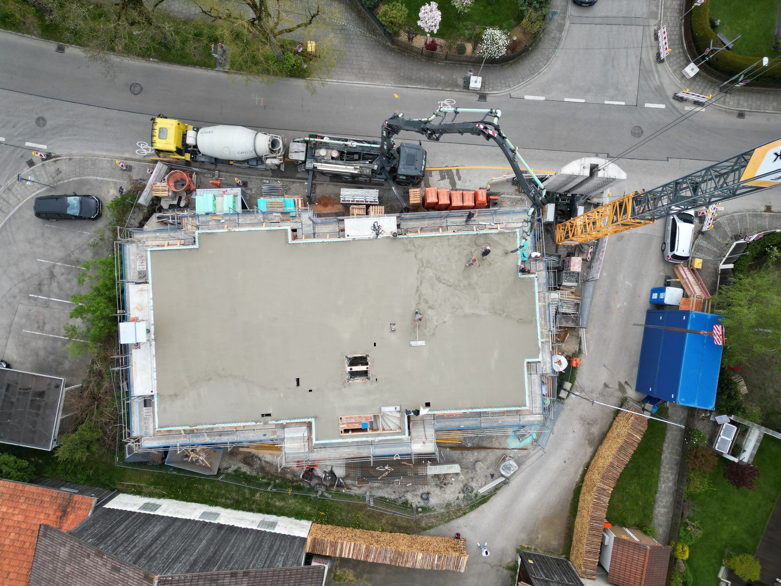 An aerial view of a construction site on a building, with construction equipment, scaffolding, and workers present. Adjacent streets and surrounding residential area are visible.