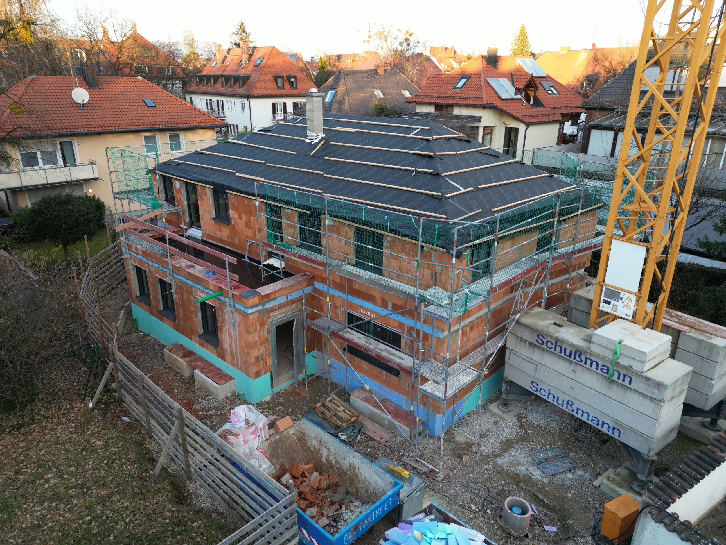 A house under construction with scaffolding around it, showing brick walls and a black roof, in a residential neighborhood.