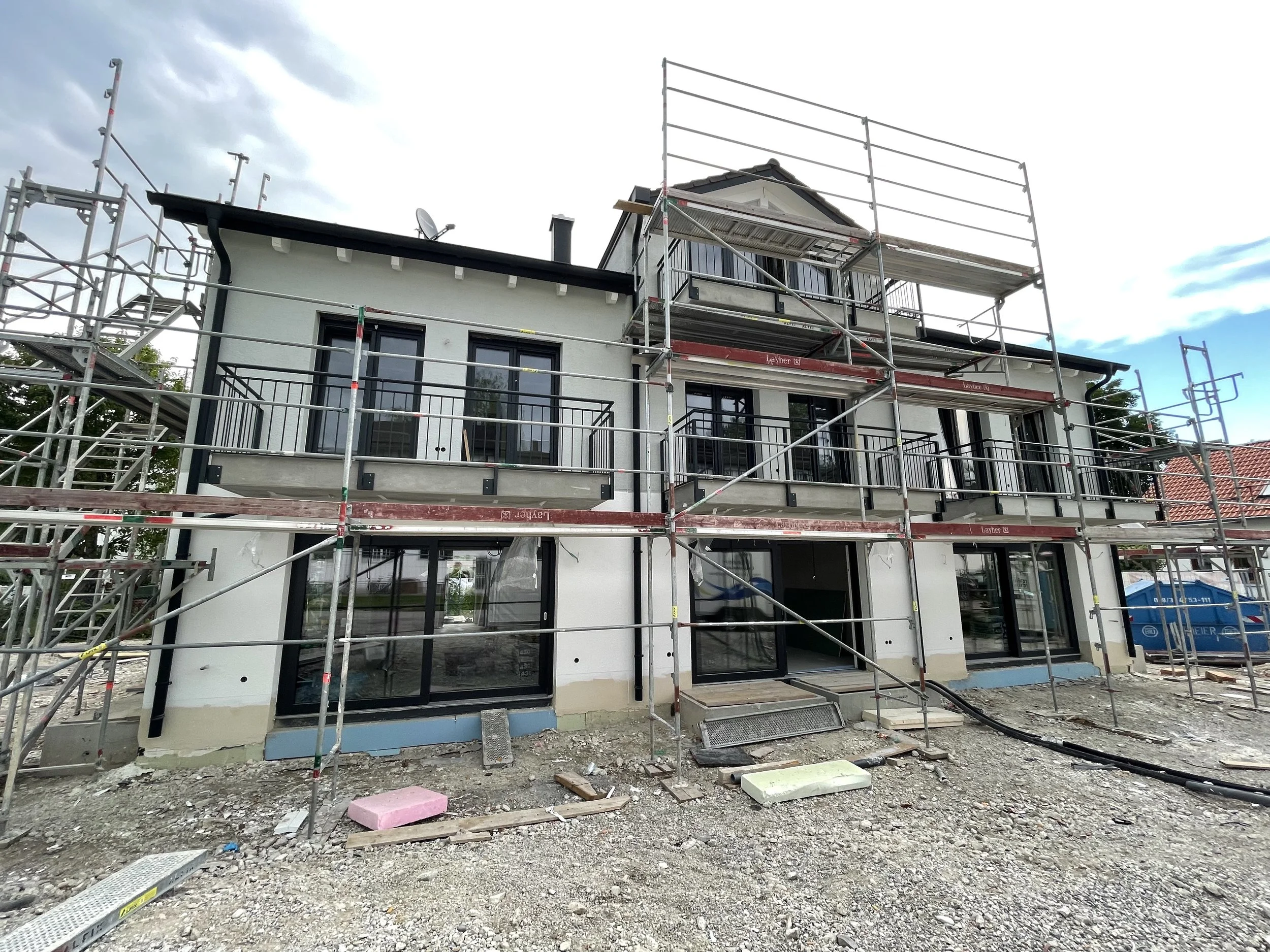 A two-story house under construction with scaffolding surrounding it, multiple large windows, and a sloped roof. The construction site has scattered construction materials and debris in the foreground.