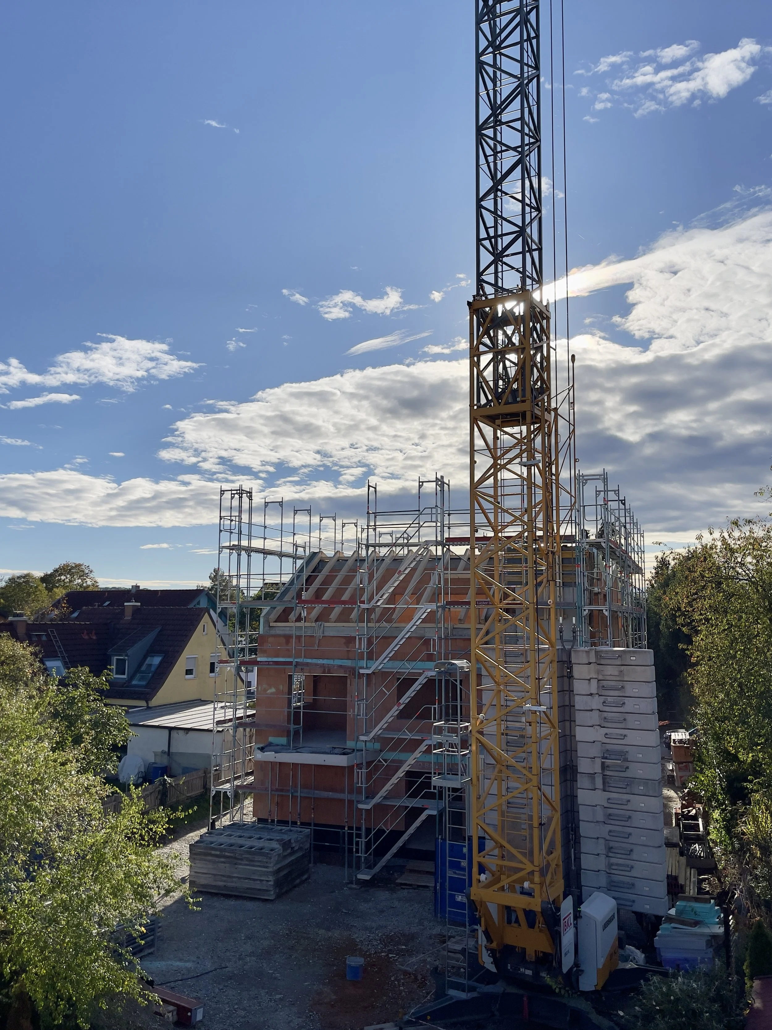 A construction site with a yellow tower crane next to a partially built house with scaffolding, surrounded by trees and neighboring houses under a partly cloudy sky.