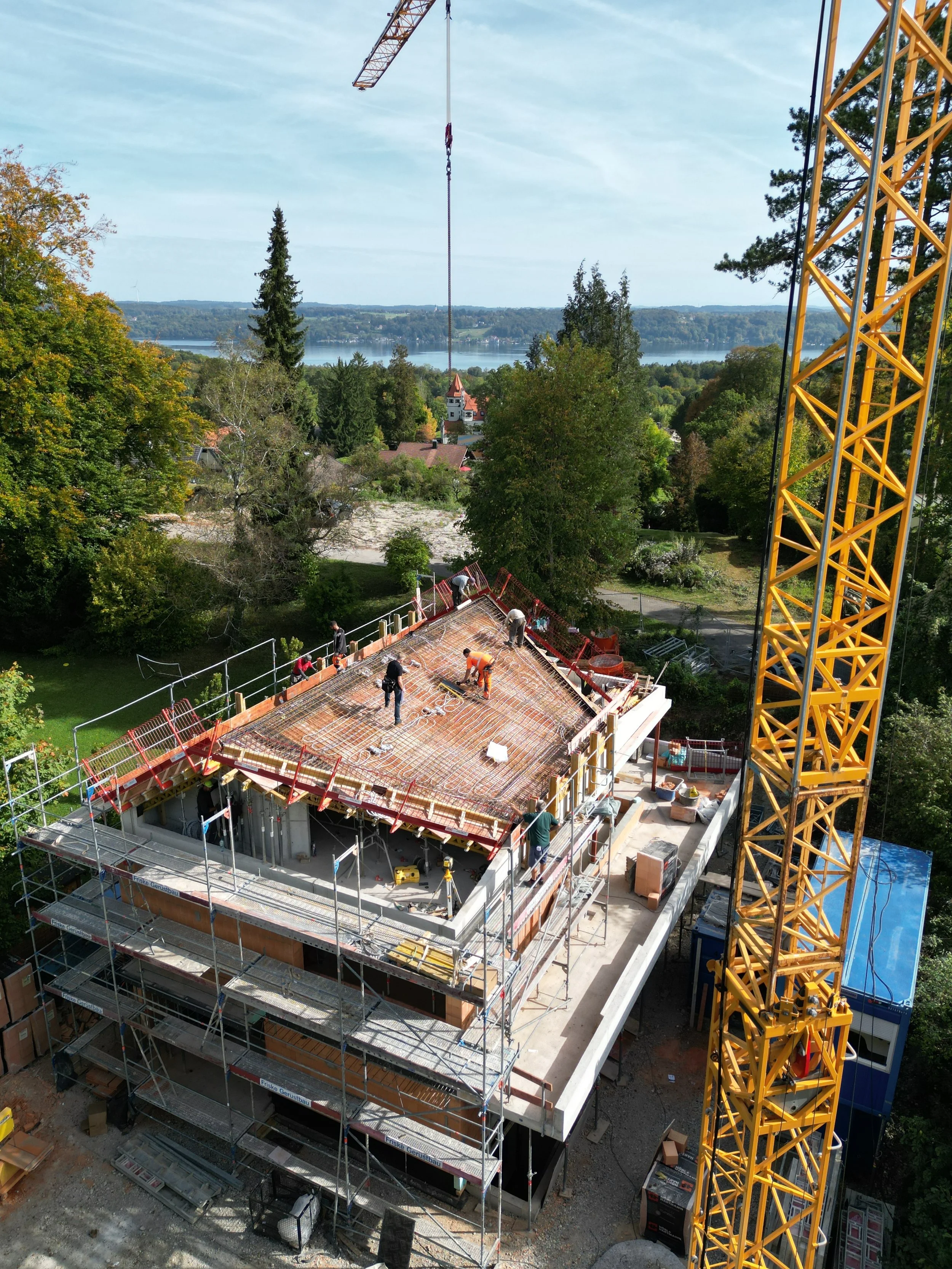 Construction site with workers building a multi-story structure, a large yellow crane on the right side, and a scenic view of trees and water in the background.