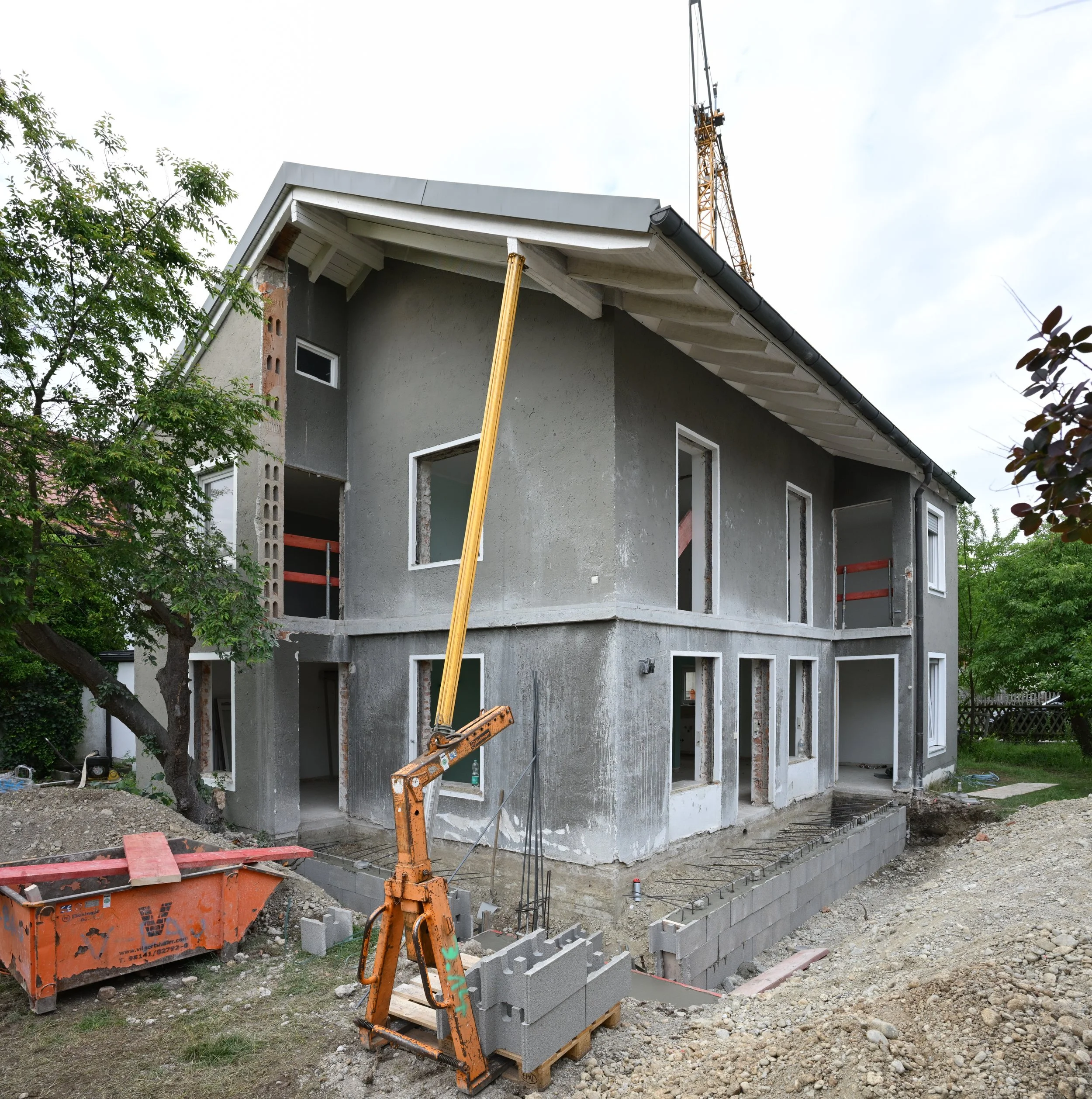 Under construction house with partially built gray walls, open windows, and construction equipment, including a yellow support pole and an orange cement mixer, in a yard with trees and gravel.