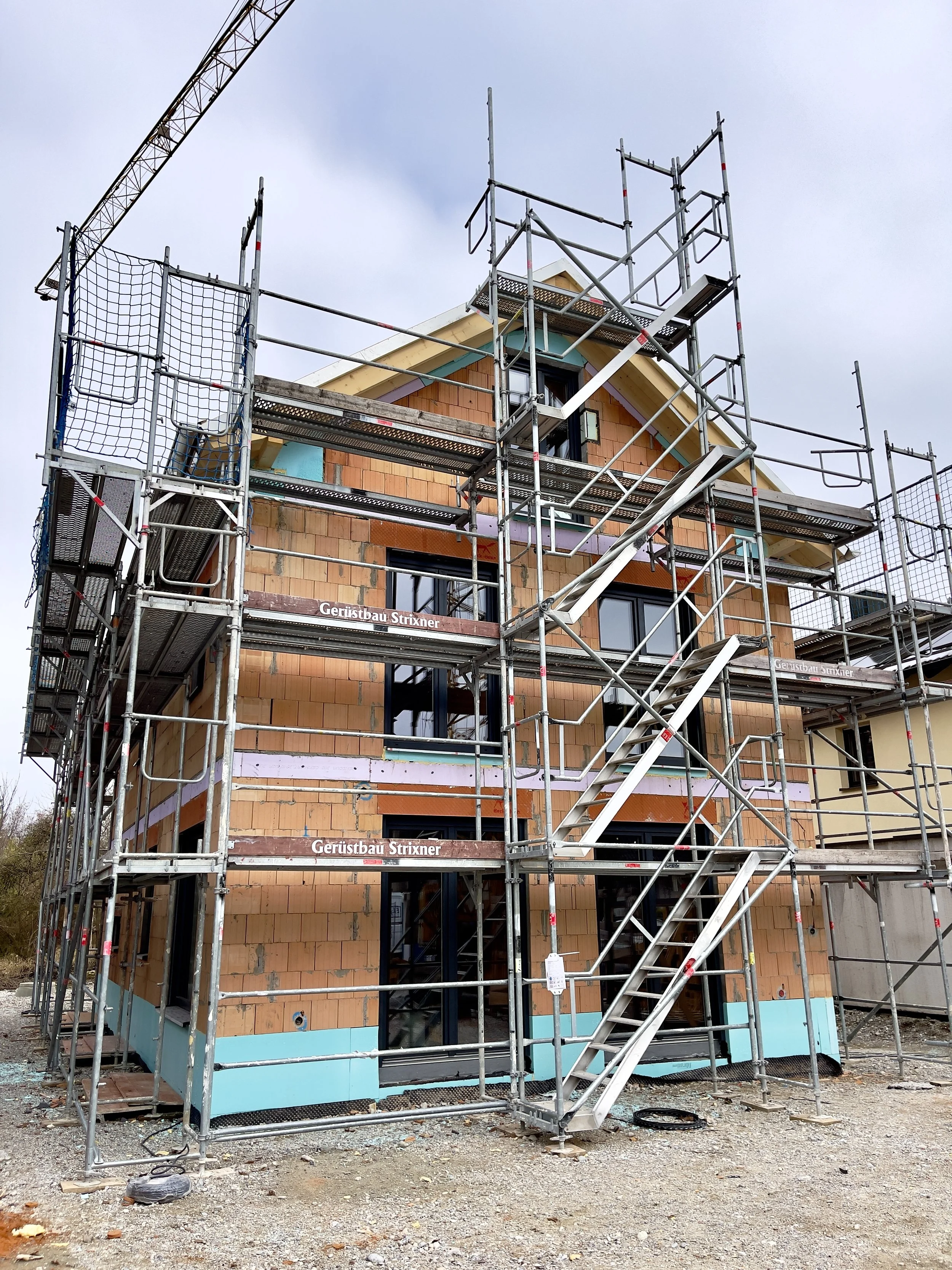 Construction site with scaffolding around a multi-story house under construction, brick walls, windows, and a crane in the background.