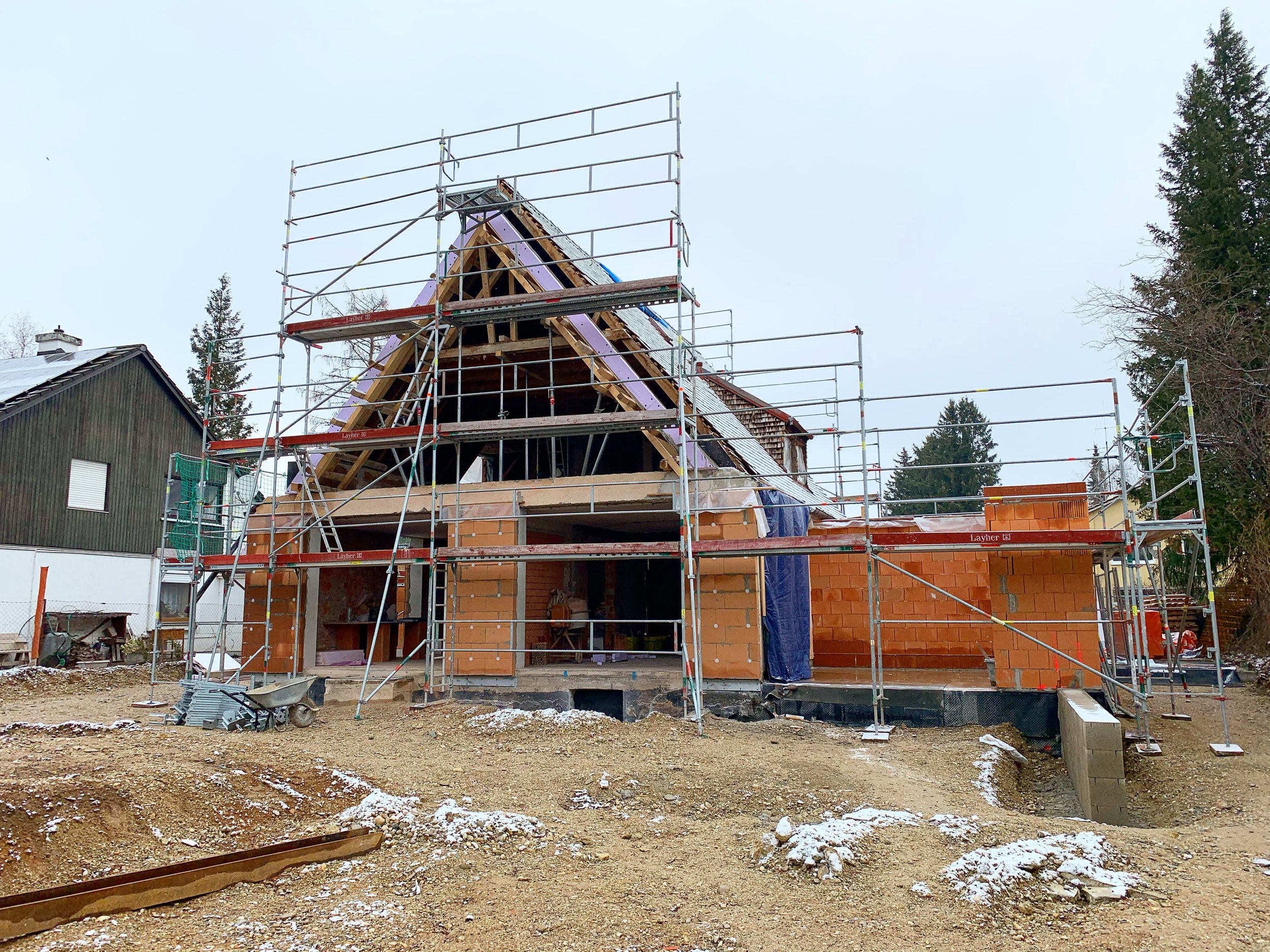 Construction site with a house in early stages of building, surrounded by scaffolding, with some snow on the ground and trees in the background.