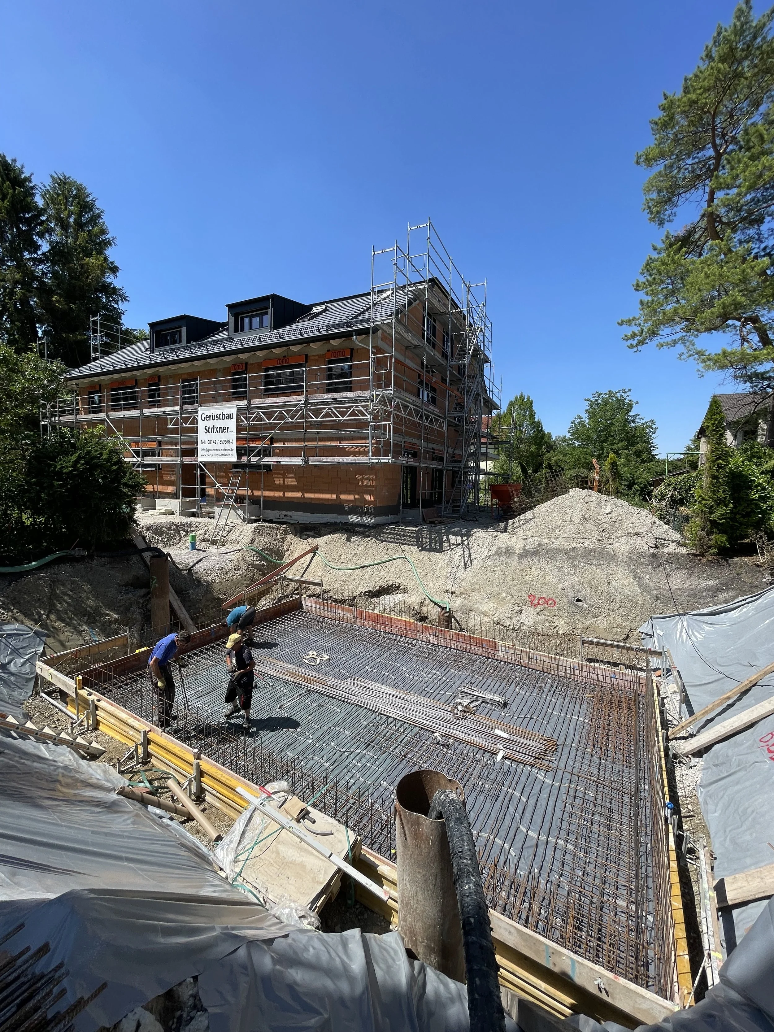 Construction site with a building under construction, workers working on concrete reinforcement, scaffolding around the building, and construction equipment on site during daytime with trees and a blue sky in the background.