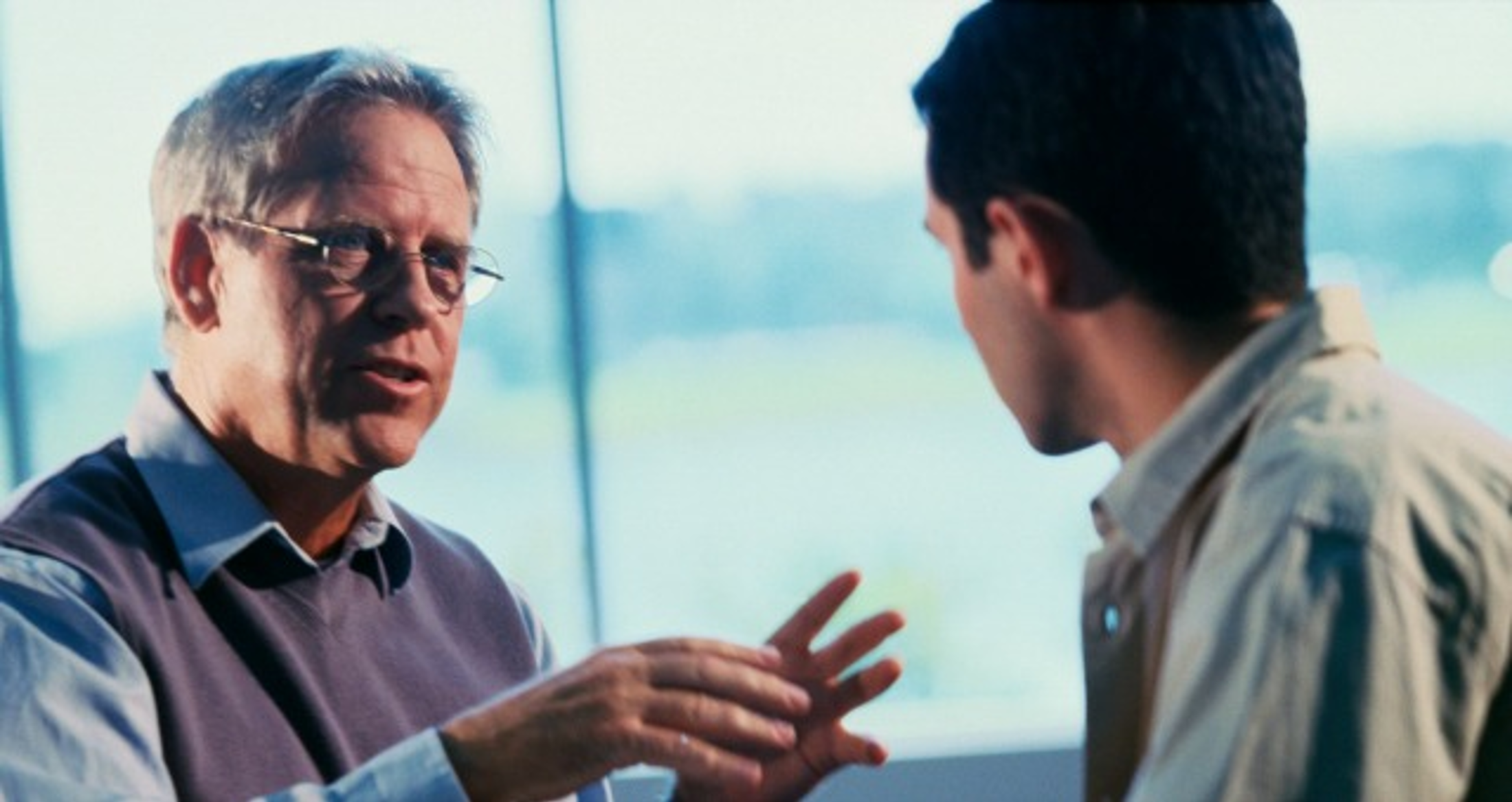 Two men are engaged in conversation indoors with large windows behind them.