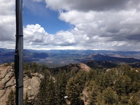 Panoramic view of mountain landscape with blue skies, scattered clouds, rocky terrain, and dense forest from a high vantage point.
