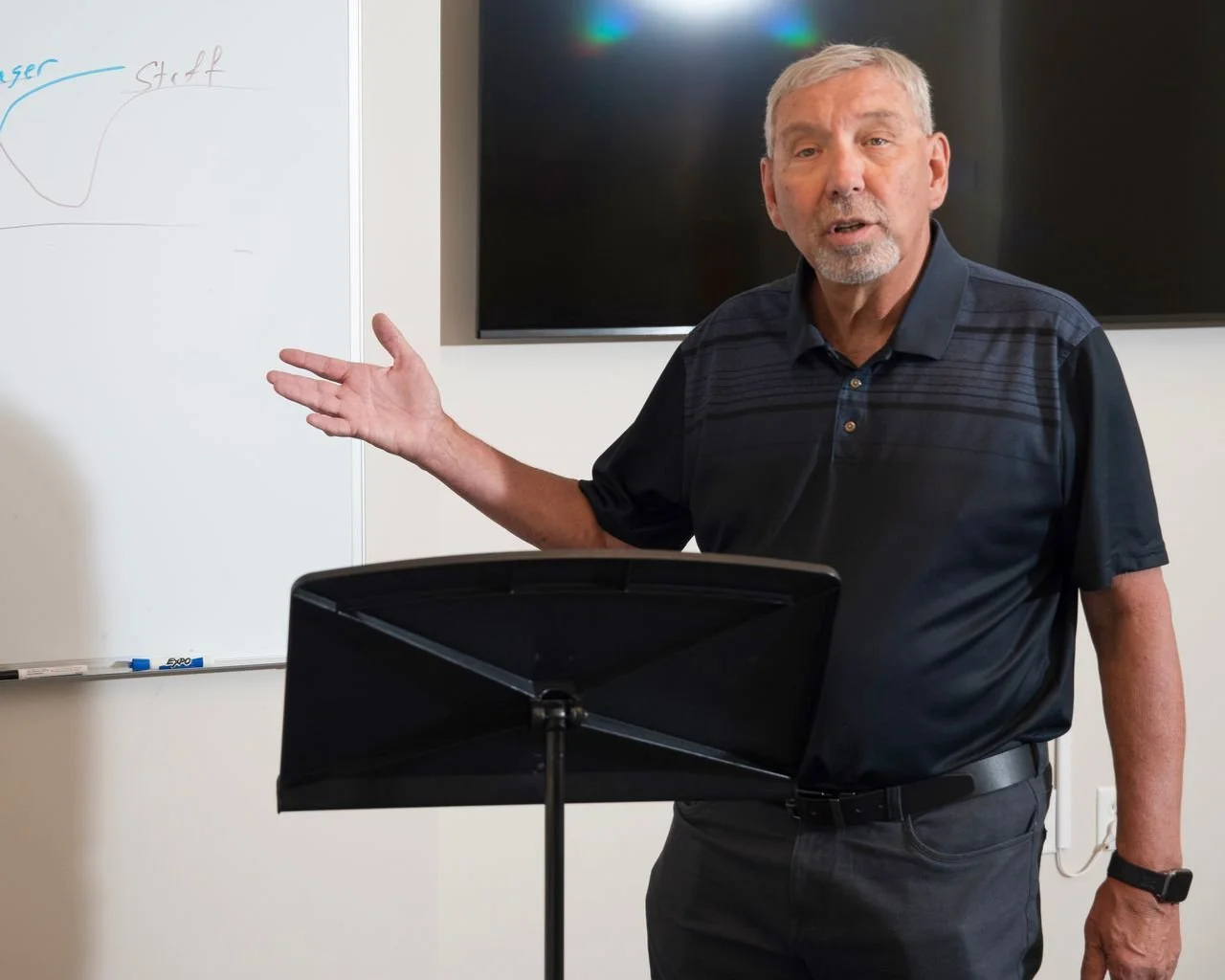 A man with gray hair and a beard, wearing a black polo shirt, is standing in front of a whiteboard with a diagram. He is gesturing with his left hand and speaking, possibly giving a presentation or lecture.