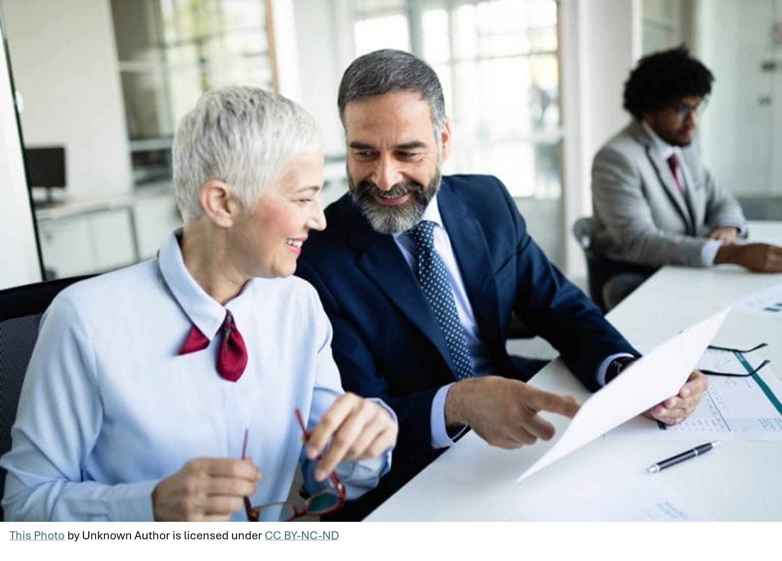 Two professionals in business attire smiling and looking at papers together in a modern office conference room, with a third person in the background working at a desk.