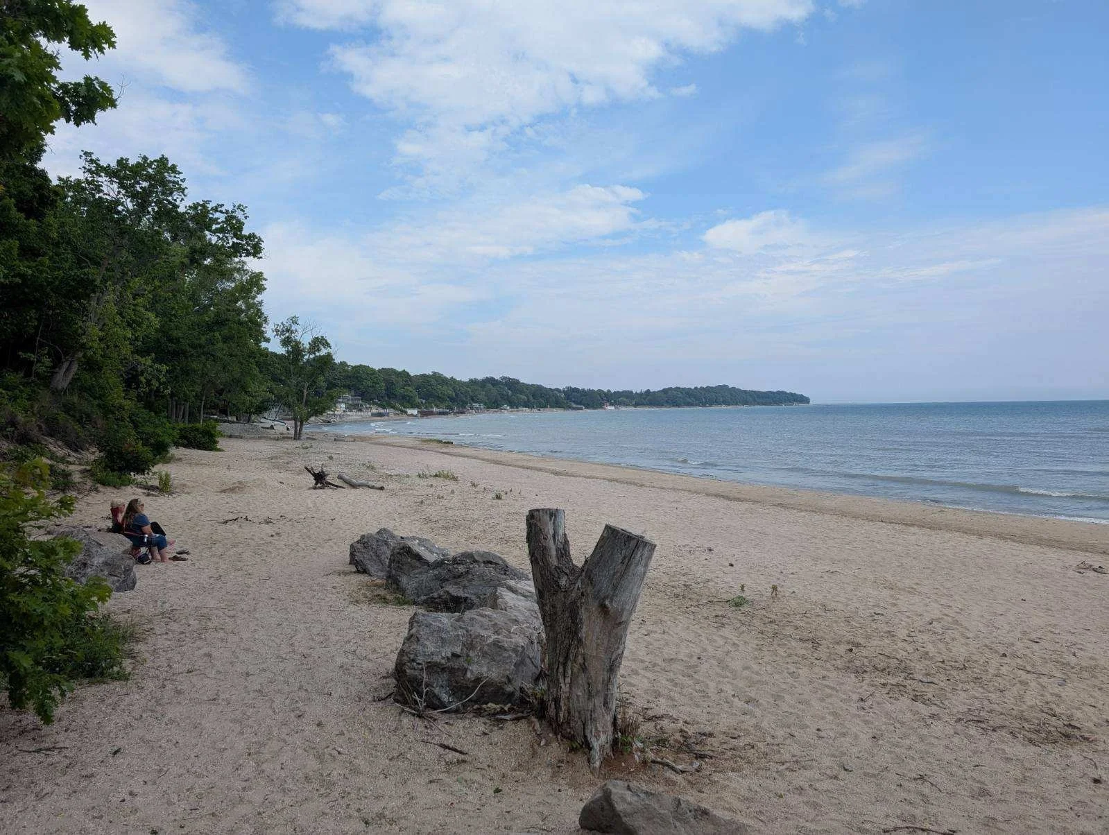 A peaceful beach scene with a sandy shoreline, green trees on the left, and calm water extending to the horizon under a partly cloudy sky.