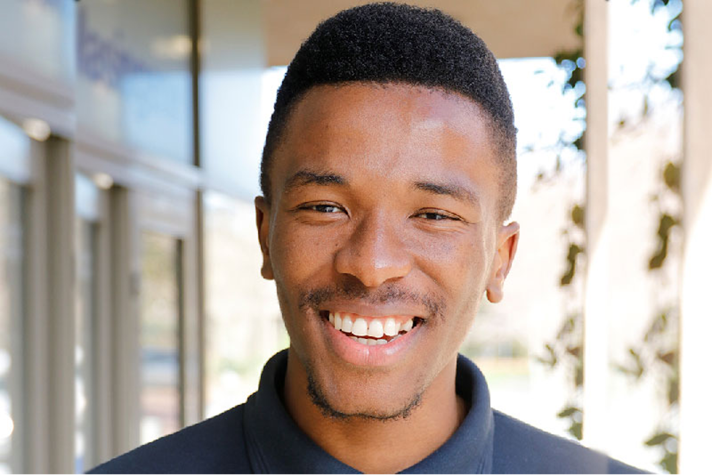 Close-up of a young man smiling with white teeth outdoors, blurred background with trees and a modern building.