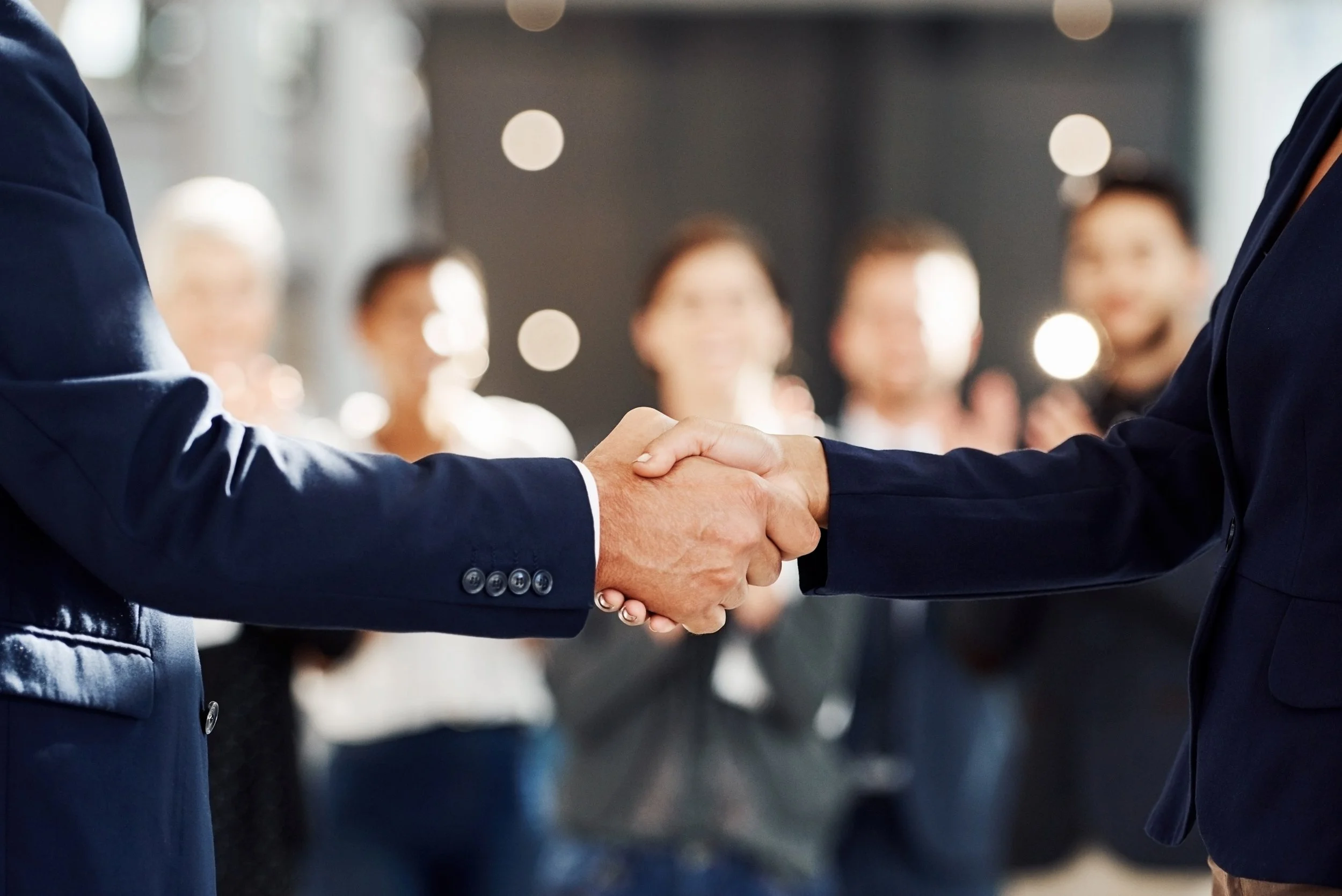 Close-up of two businessmen in suits shaking hands, with a blurred group of professionals in the background.