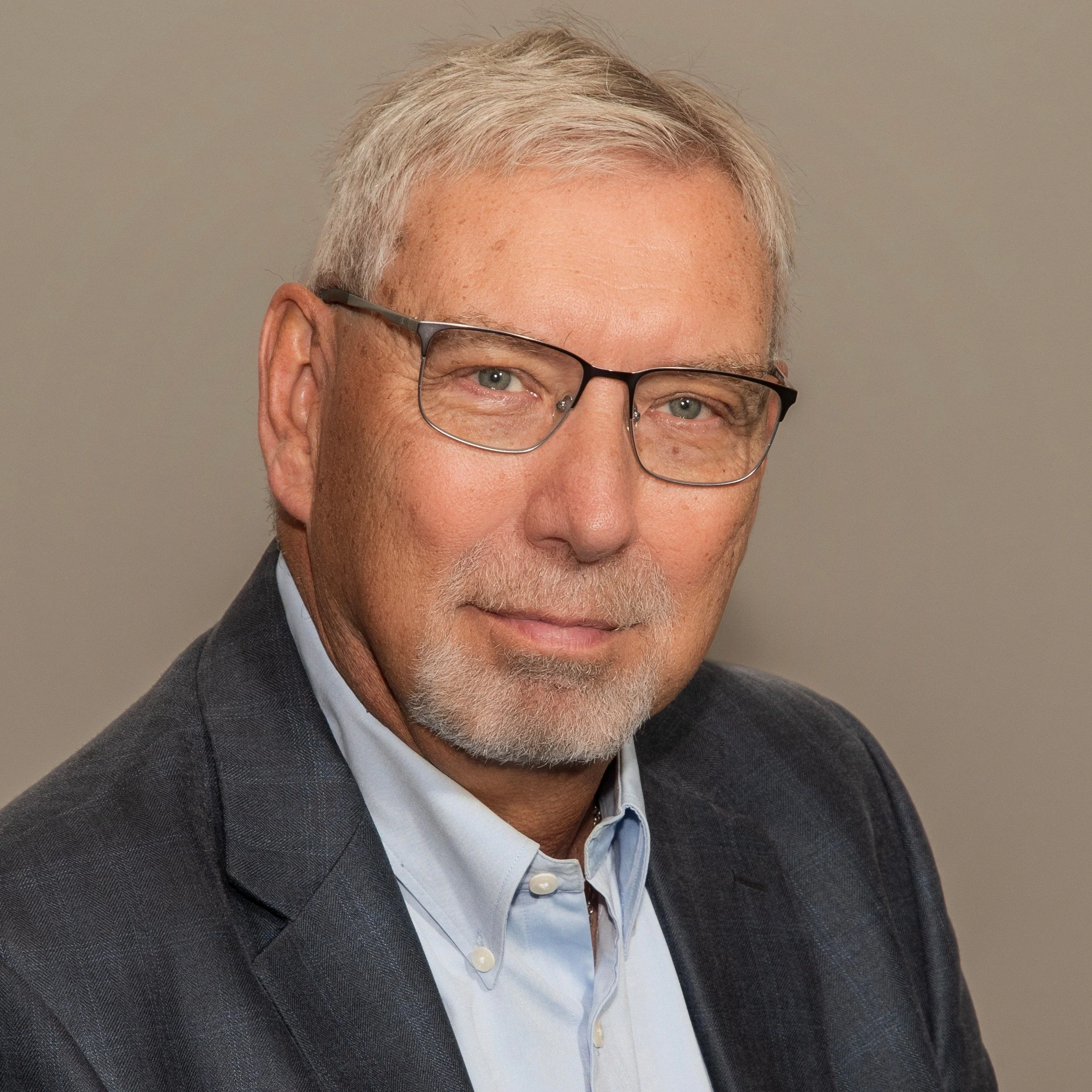 Close-up of a middle-aged man with gray hair and beard, wearing glasses, a dark gray suit, and a light blue shirt, looking directly at the camera against a plain background.