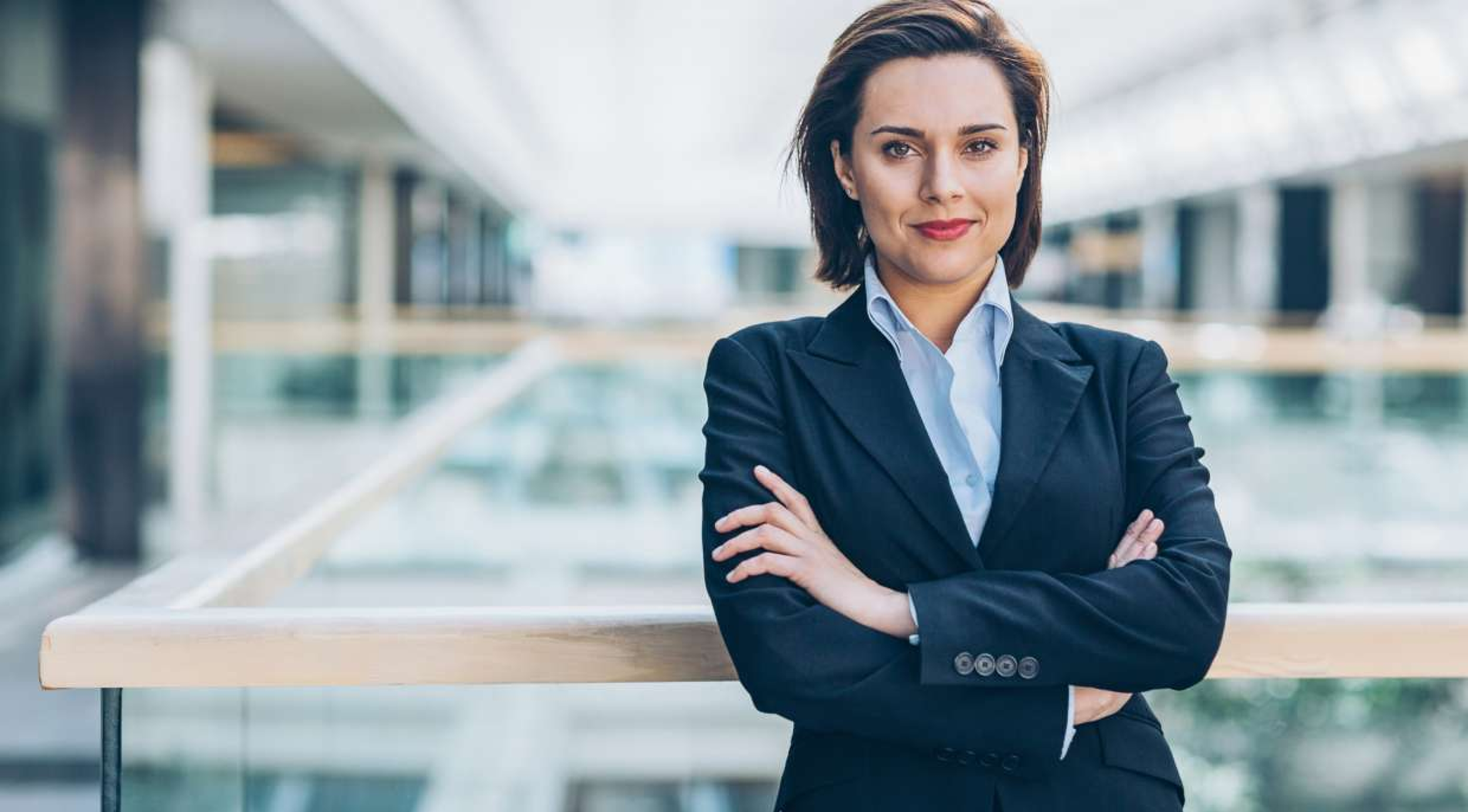 A businesswoman with short brown hair and a confident smile, wearing a dark blazer and light blue shirt, standing with crossed arms near a railing in a modern office or airport building with glass and metal architecture.