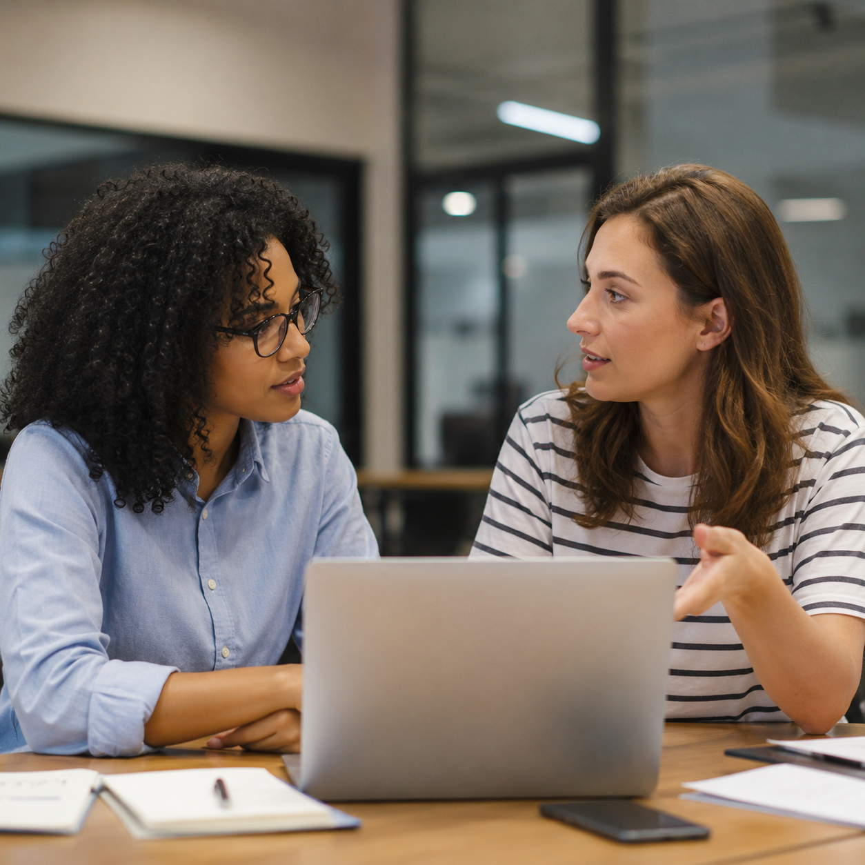 Two women having a discussion at a table with a laptop, notebooks, and a smartphone, in a modern office setting.