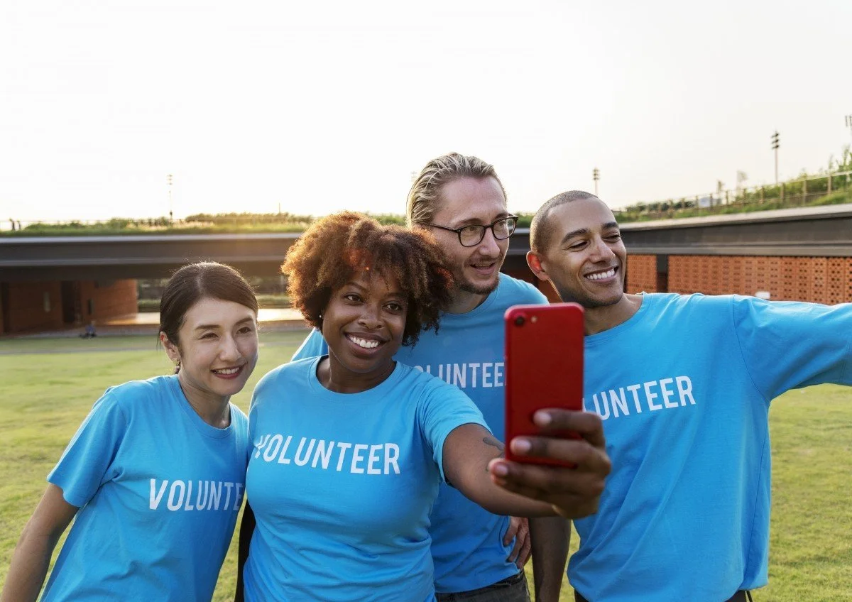 Four volunteers in blue shirts with 'Volunteer' written on them, taking a group selfie outdoors on a grassy field during daytime.
