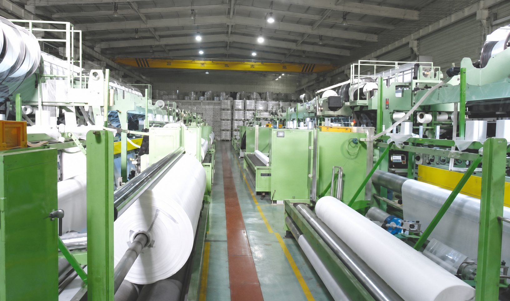 Factory interior with green machinery and large rolls of white material, possibly a manufacturing or packaging facility.