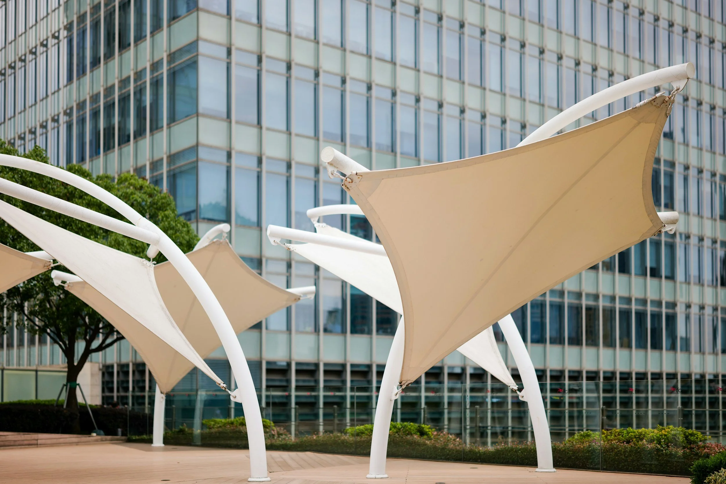 Multiple white shade structures in front of a modern glass building with reflective windows, greenery, and pathway.