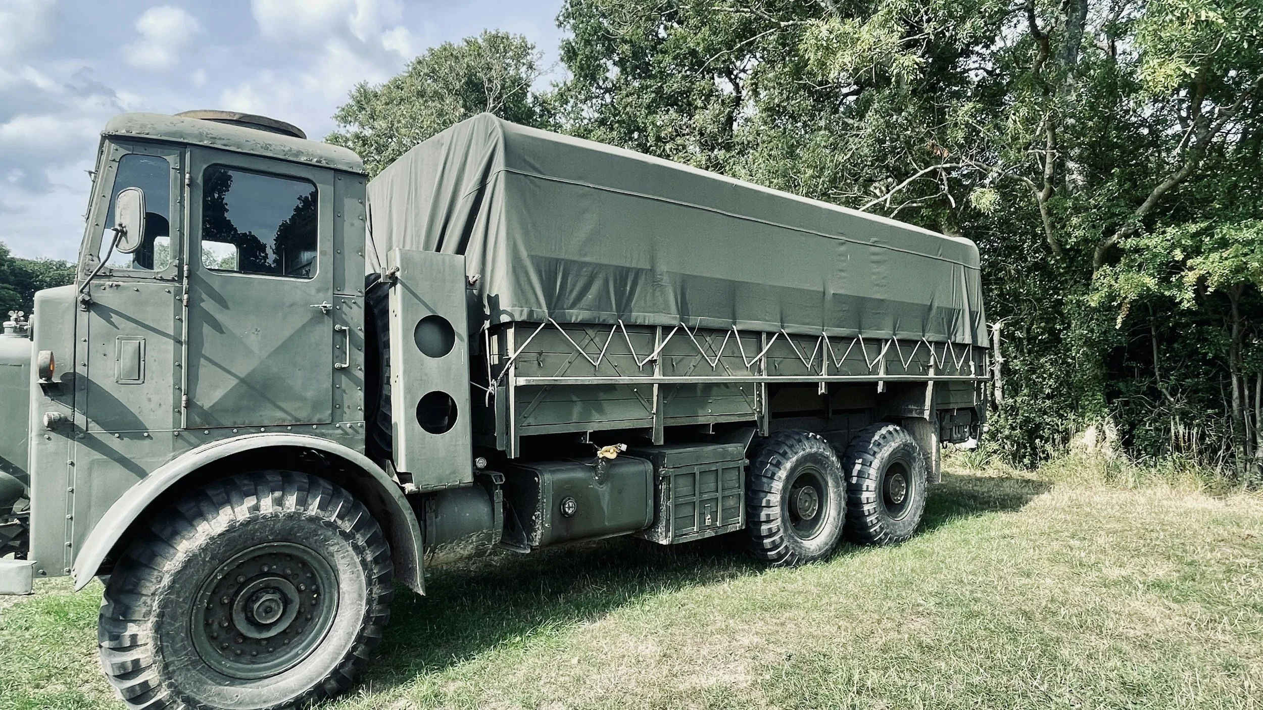A military green truck with a covered cargo area parked on grass near trees.