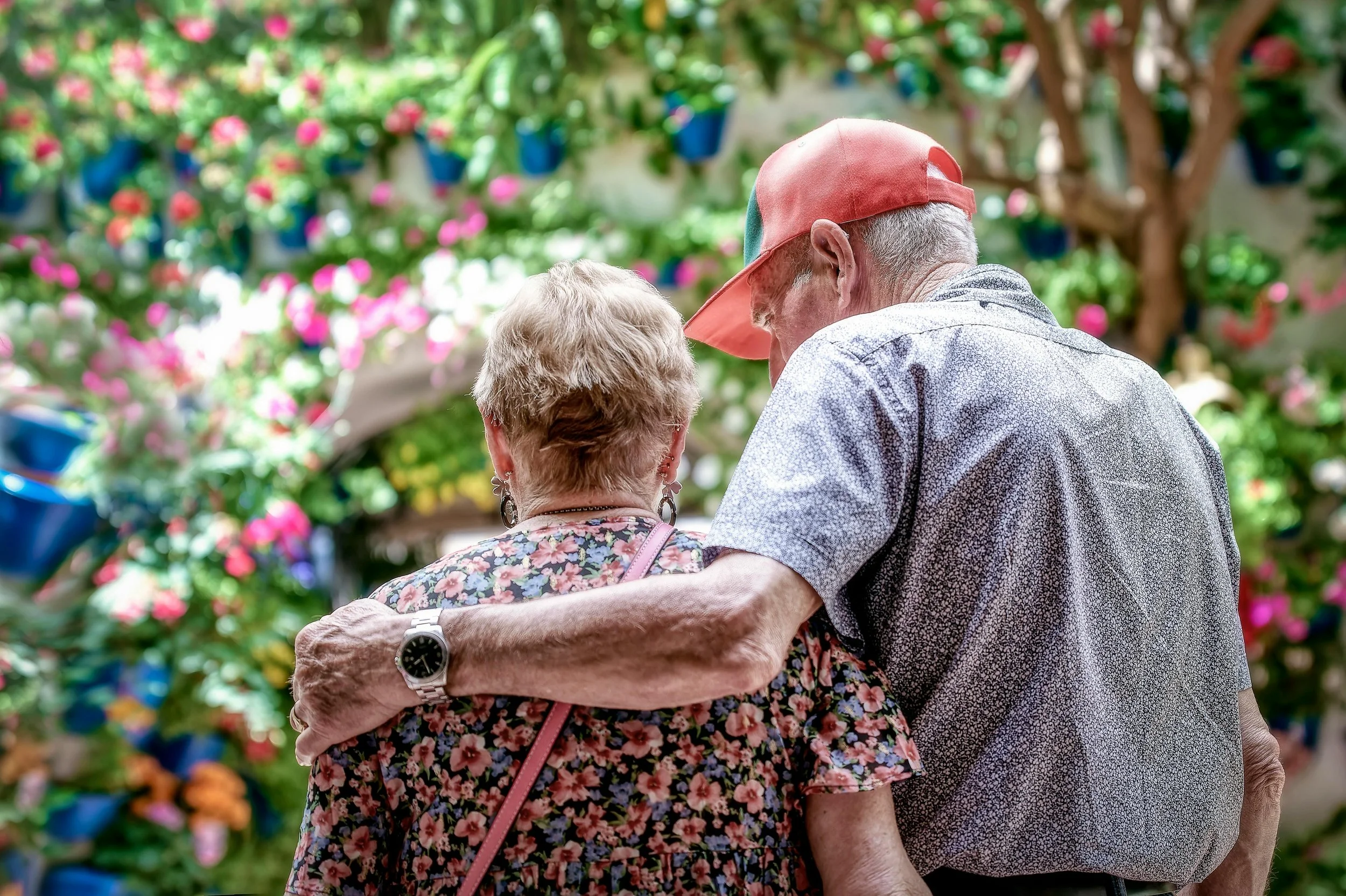 An elderly man and woman standing close together, embracing in a garden with pink and red flowers and hanging blue flower pots.