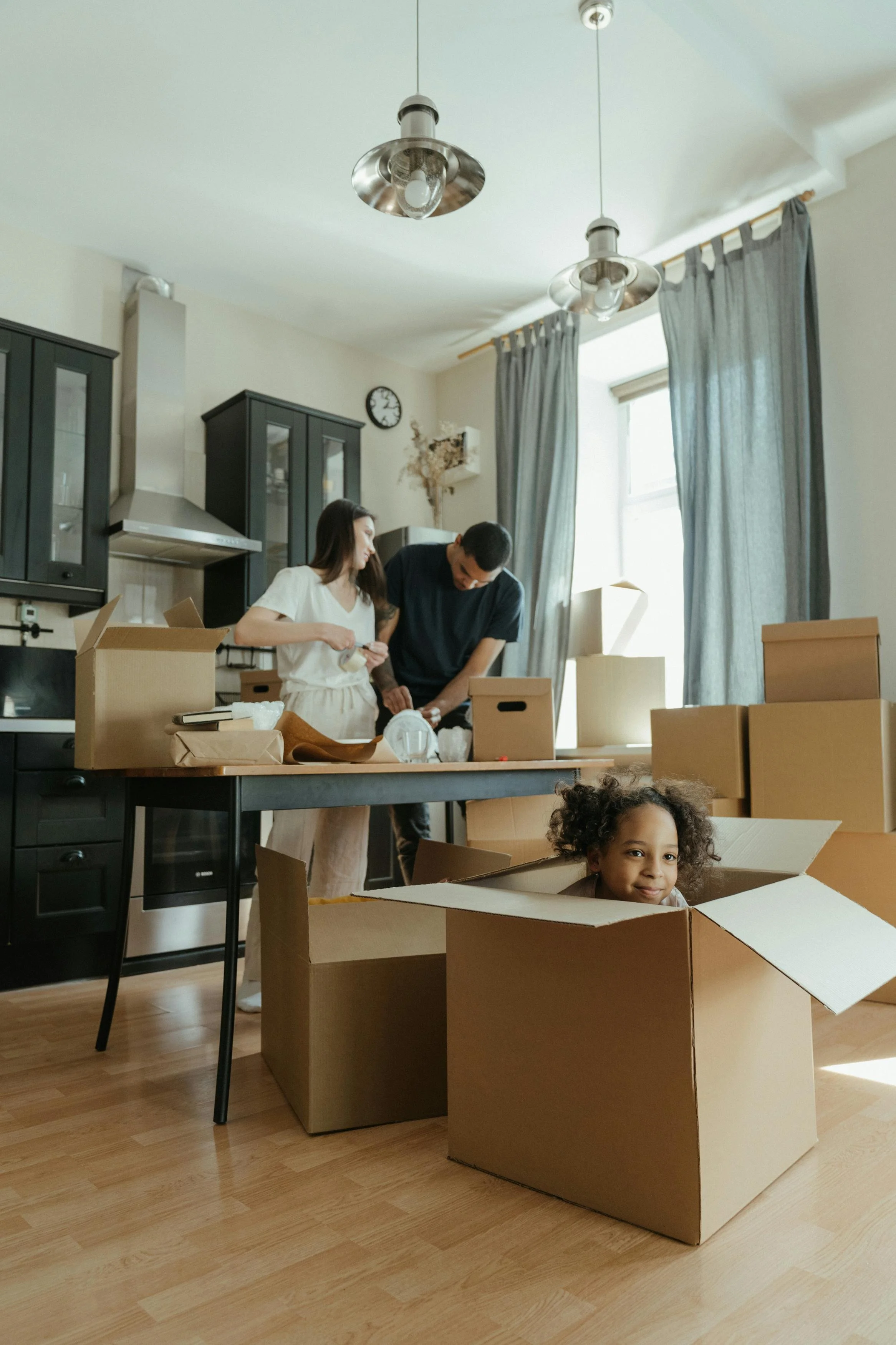 Two adults and a child are unpacking moving boxes in a bright kitchen with black cabinets and gray curtains. The child is sitting inside a large cardboard box, smiling at the camera. The adults are working together, with one woman holding a small box and a man leaning over a box on the table. The scene has natural sunlight coming through a window.