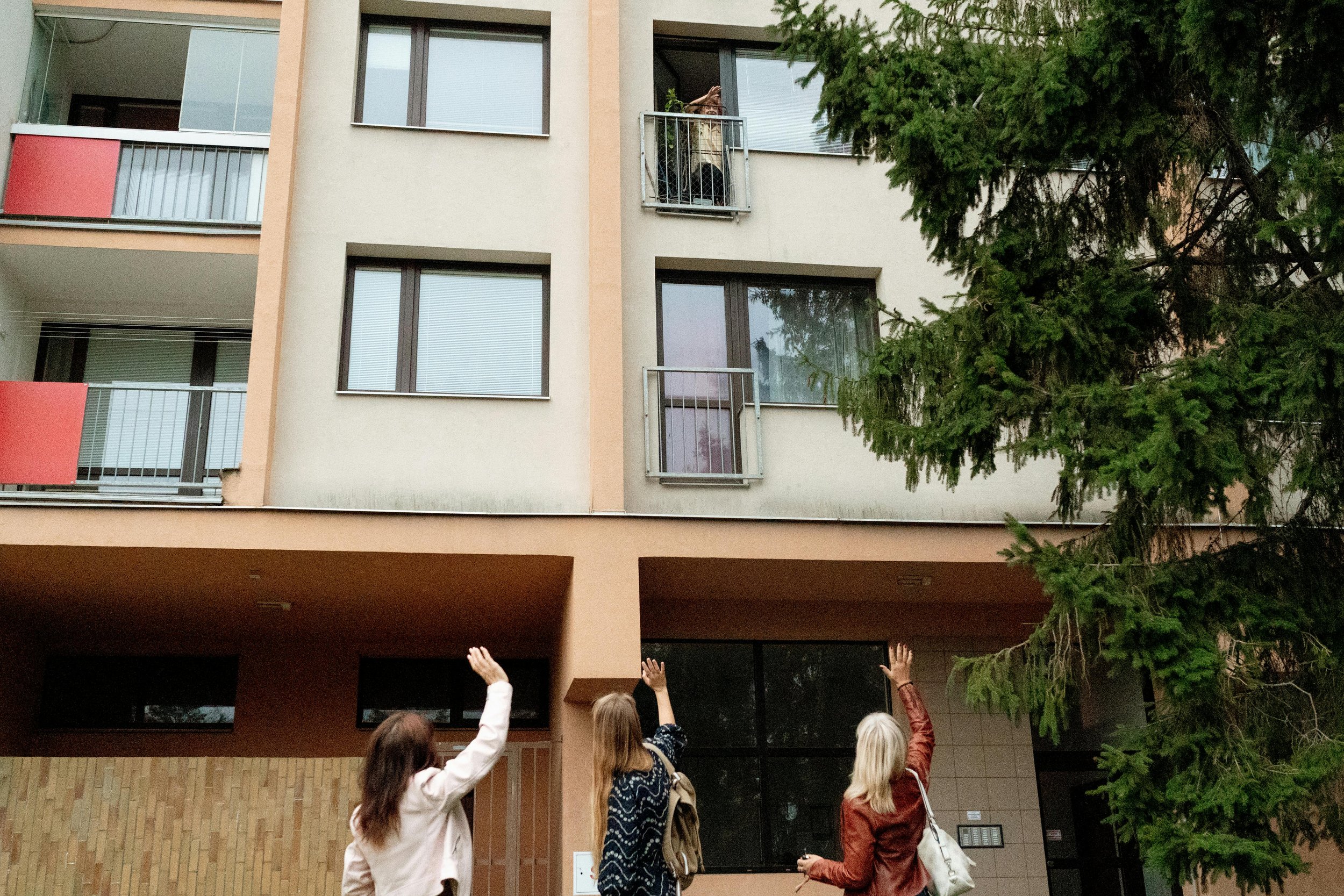 Three people with their backs facing the camera and waving towards a person standing on a balcony of an apartment building.