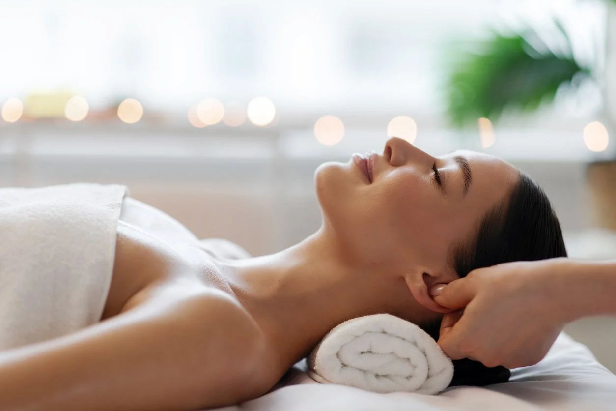 Woman receiving a head massage in a spa or wellness setting, lying on a massage bed with a rolled towel under her neck, relaxing with eyes closed.