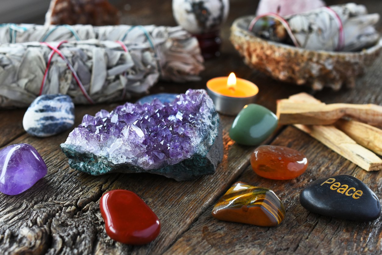 Collection of colorful gemstones, crystals, and candles arranged on a rustic wooden surface, with some stones inscribed with words like 'Peace' and 'Love'.