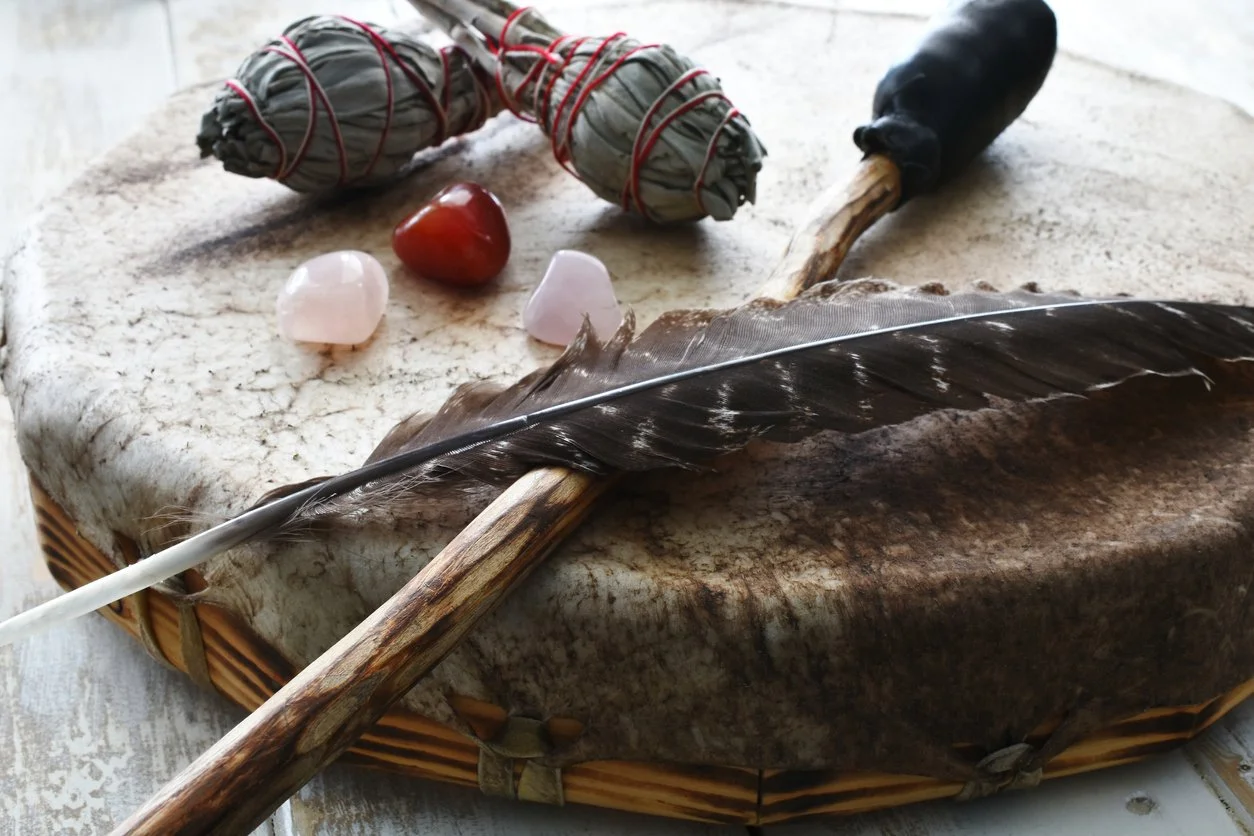 A collection of mystical items on a stone surface includes two sage smudge bundles tied with red string, a black feather quill with a wooden handle, a small round red stone, and two pink stones, all arranged on a large woven tray.
