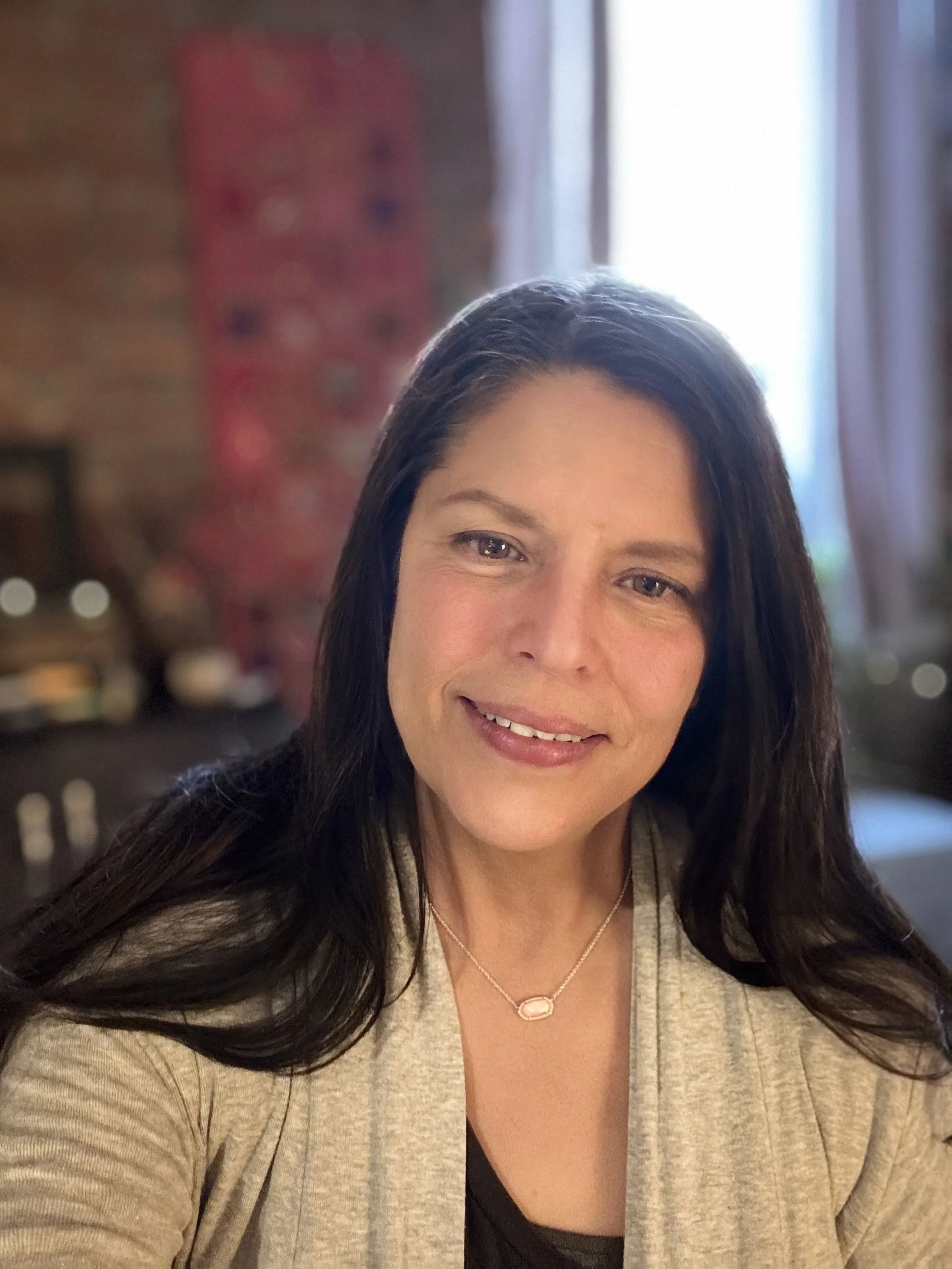 Close-up of a woman with long dark hair, smiling, wearing a beige cardigan over a black top, and a delicate necklace, with a blurred indoor background.