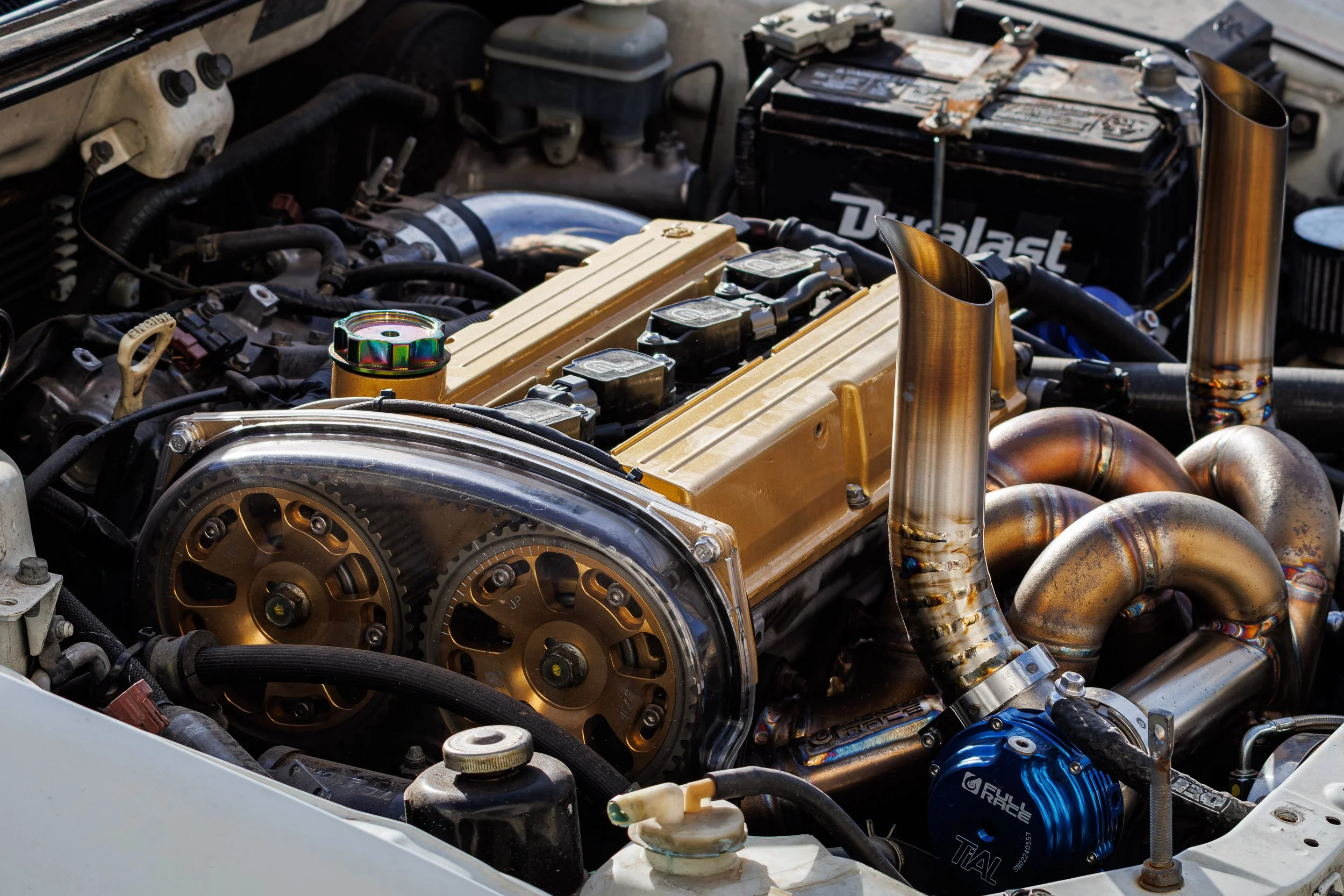 Detail car photography showcasing the heavily modified engine bay of a JDM Mitsubishi Lancer Evolution, featuring exposed cam gears, a custom hood-exit exhaust, and a Full-Race turbo setup in Scottsdale, Arizona, captured by Curtis from Gasoline and 