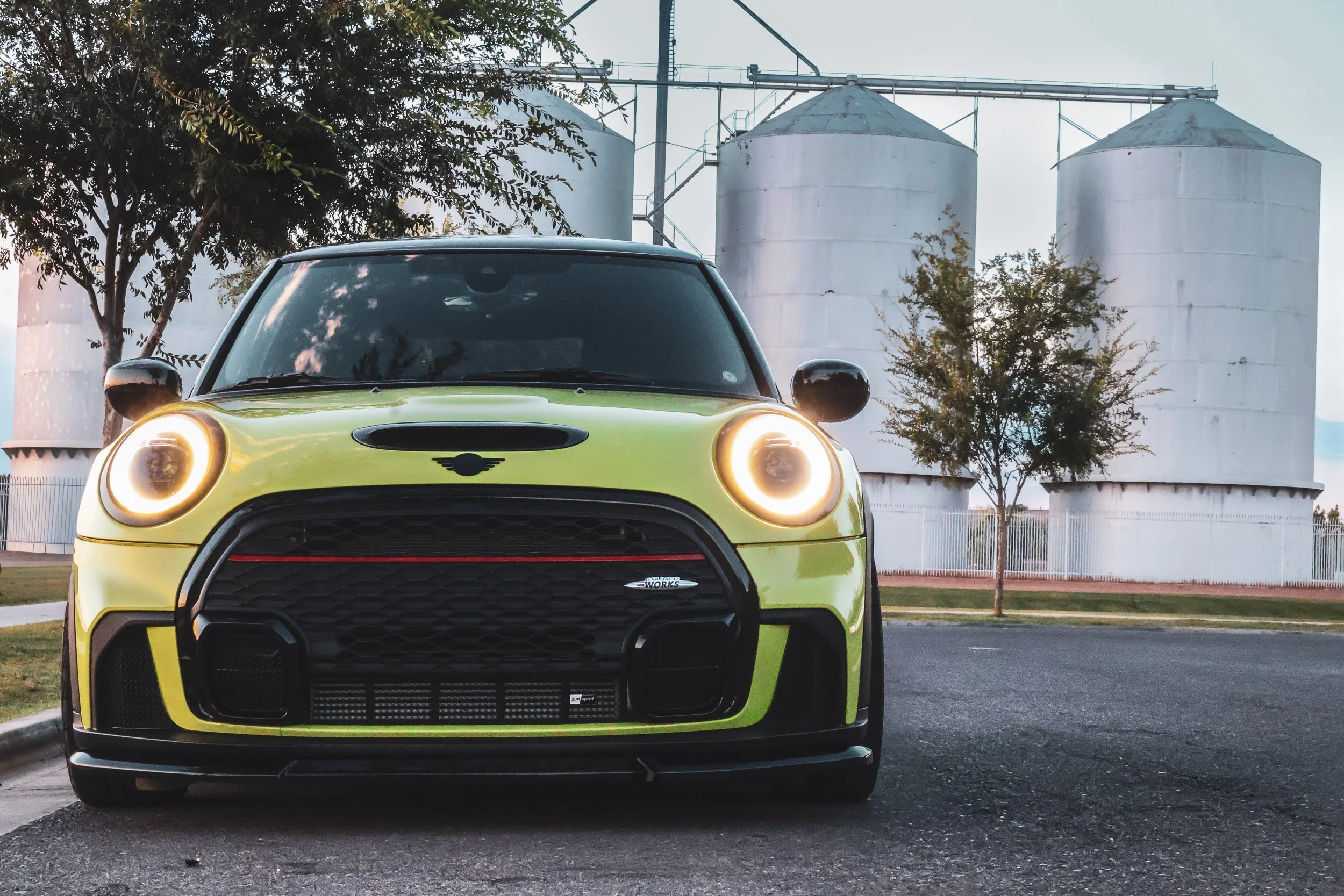 Low-angle front-facing automotive photography showcasing a vibrant yellow Mini Cooper JCW with glowing halo headlights, parked in front of the historic Morrison Ranch silos at dusk in Gilbert, Arizona, photoshoot by Curtis at Gasoline and Dopamine.