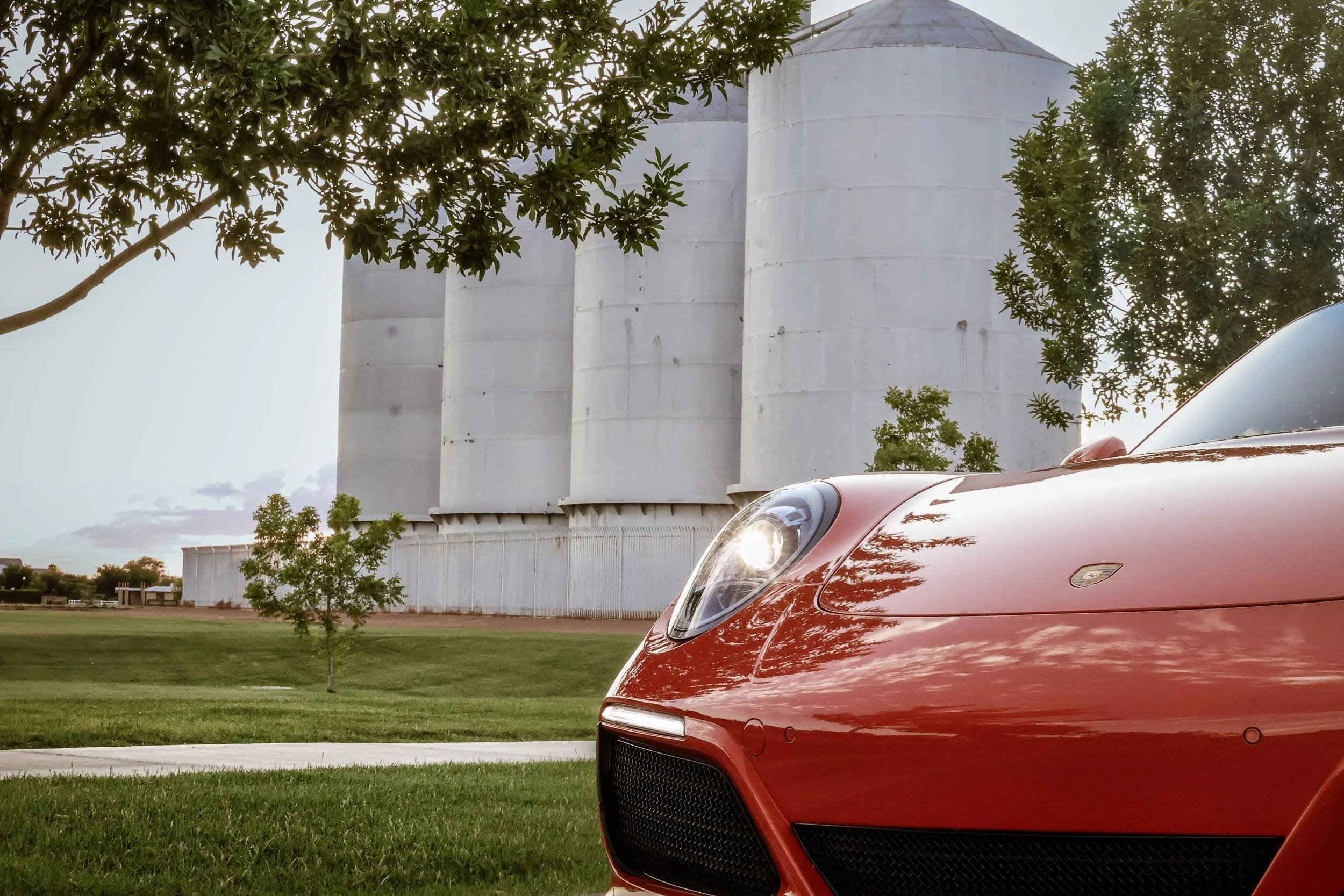 Close-up automotive photography focusing on the sleek hood and illuminated headlight of a Lava Orange Porsche 911 C4S, framed against the historic Morrison Ranch silos in Gilbert, Arizona, captured by Gasoline and Dopamine.