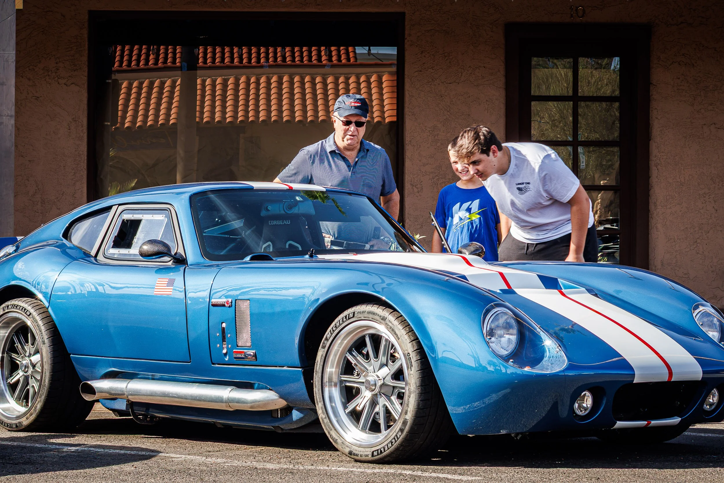 Candid automotive lifestyle photography of three generations of car enthusiasts admiring the interior of a classic blue and white striped Shelby Cobra on a sunny day in Arizona, captured by Curtis from Gasoline and Dopamine Car Photography in Scottsd