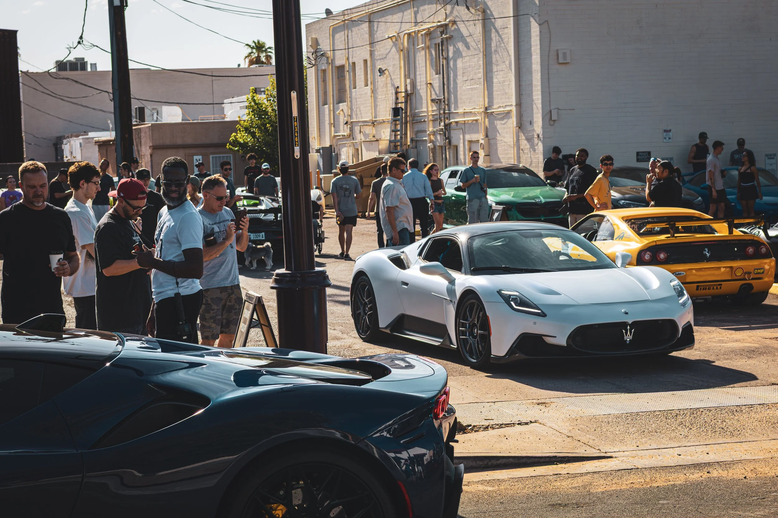 A lovely wide shot of a car meet with a number of colorful supercars, some parked and some driving, with many people nearby watching and admiring.