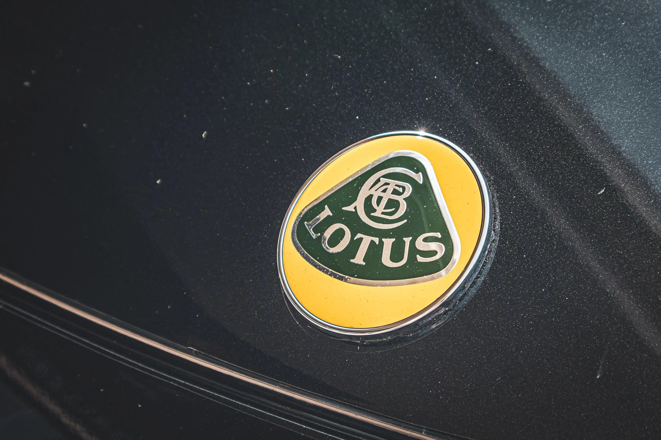 Macro detail automotive photography highlighting the iconic green and yellow Lotus emblem set against sparkling dark metallic paint in Arizona, captured by Gasoline and Dopamine.