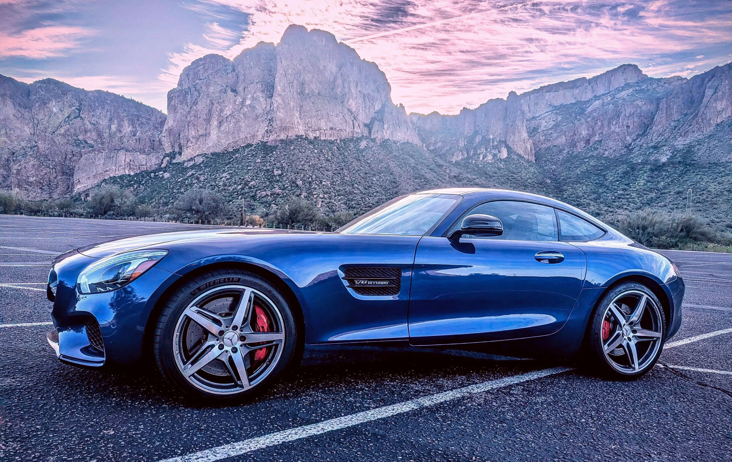 Side profile automotive photography featuring a metallic blue Mercedes-AMG GT S coupe parked in an empty lot, showcasing its long hood and sleek lines against the towering peaks of the Superstition Mountains under a colorful sunrise sky in Arizona, p