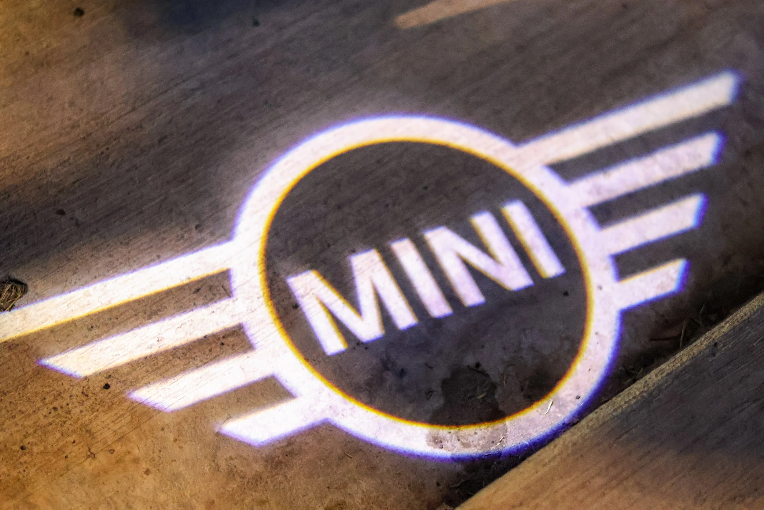 Macro automotive detail photography capturing the illuminated Mini Cooper winged logo projected onto concrete from a door puddle light at night in Morrison Ranch, Gilbert, Arizona, captured by Gasoline and Dopamine.
