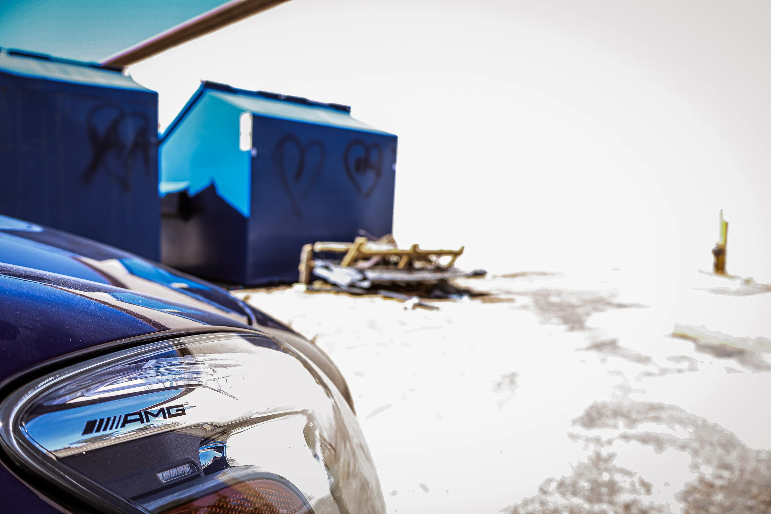 Macro automotive detail photography focusing on the etched AMG logo inside the headlight of a metallic blue Mercedes-AMG GT S, set against a highly overexposed background featuring blue industrial dumpsters tagged with spray-painted black hearts in Q