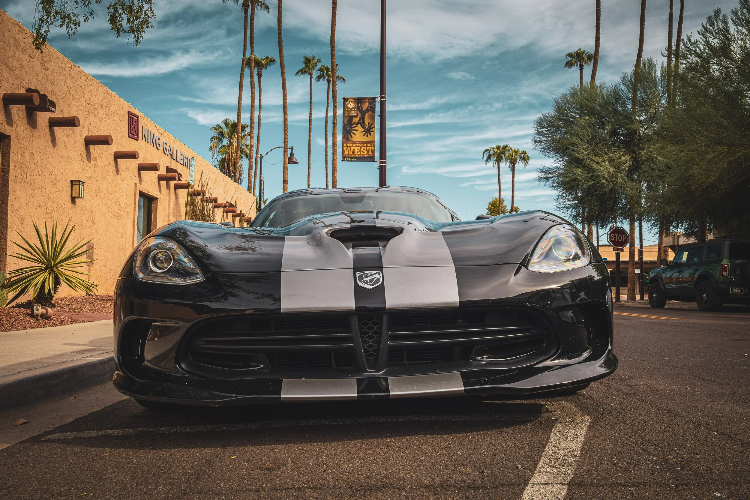 Low-angle front automotive photography highlighting the aggressive crosshair grille, hood scoop, and dual silver racing stripes of a black Dodge Viper, parked on a sunny palm-lined street in front of an adobe gallery in Scottsdale, Arizona, car photo