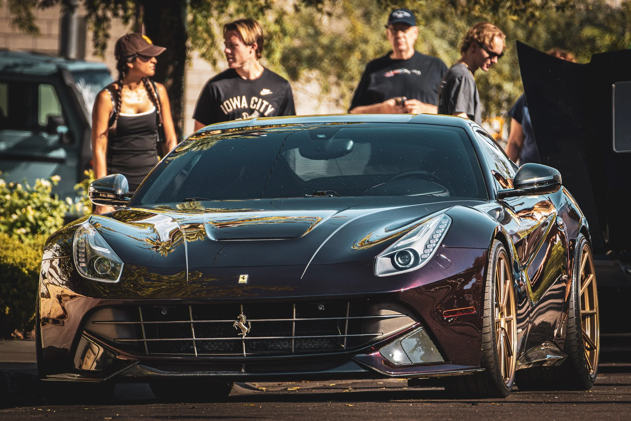 Exotic car photography capturing a low front-quarter view of a deep metallic purple Ferrari F12berlinetta featuring gold wheels and a carbon fiber front splitter, parked at a sunny cars and coffee event in Scottsdale, Arizona, captured by Curtis from