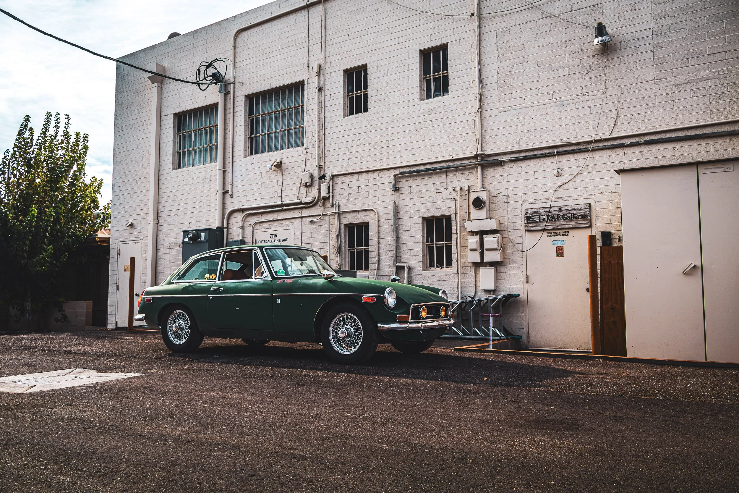 Wide-angle side profile automotive photography featuring a classic dark green MG B GT with chrome wire wheels, parked in a textured white brick alleyway behind a gallery in Scottsdale, Arizona, captured by Gasoline and Dopamine.