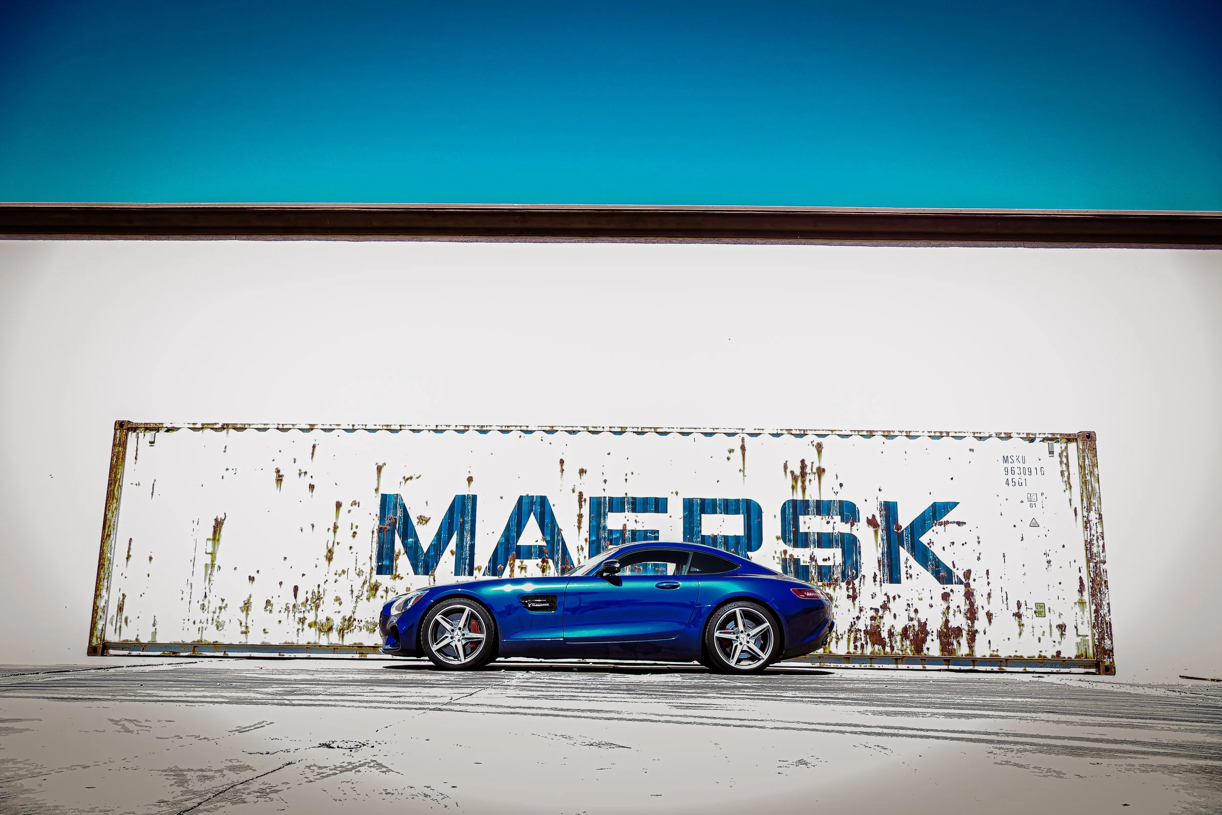 Wide-angle automotive photography capturing the sleek side profile of a metallic blue Mercedes-AMG GT S coupe, contrasting against a heavily weathered white Maersk shipping container and a stark blue sky in an industrial lot in Queen Creek, Arizona, 