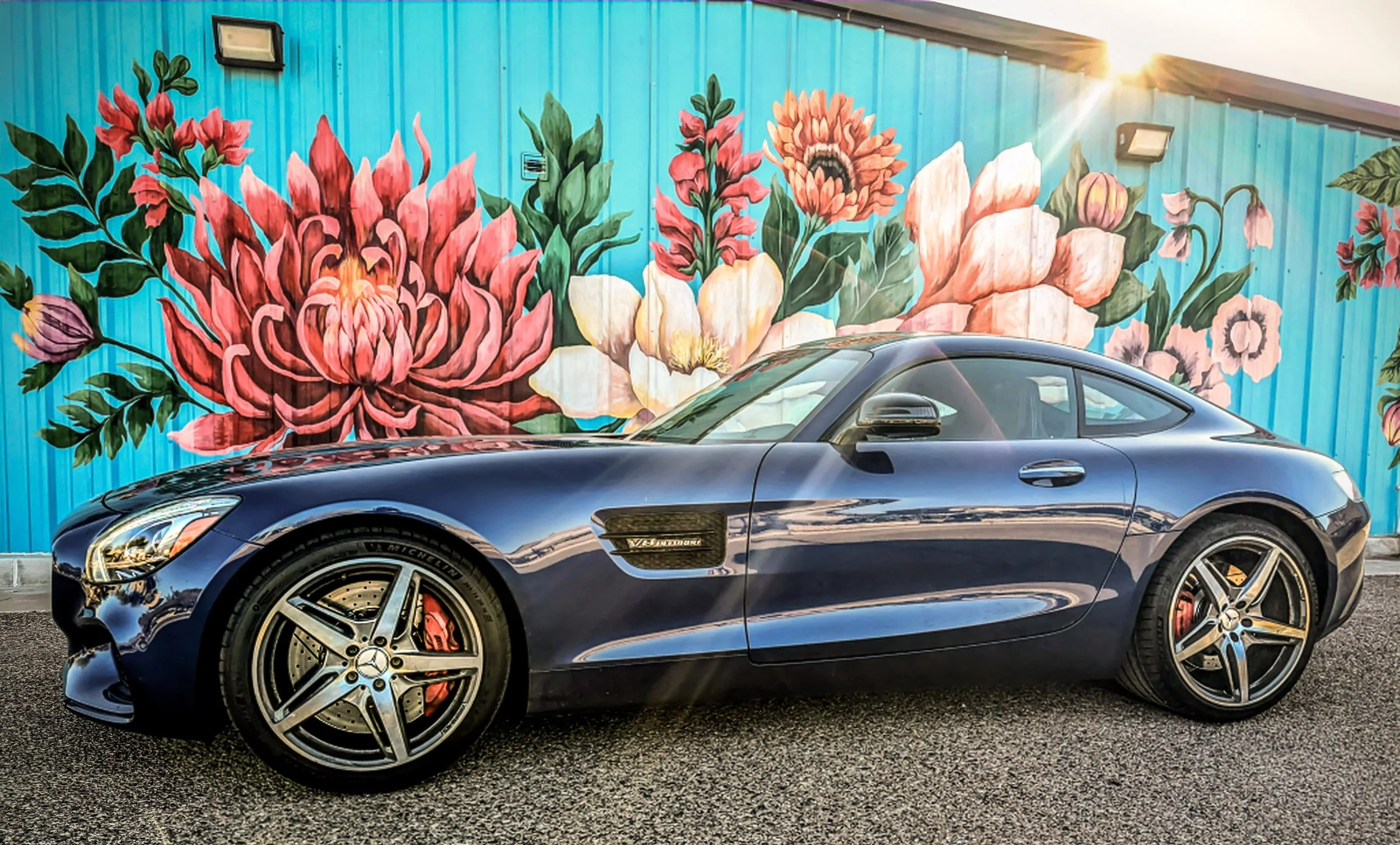 Side profile automotive photography featuring a metallic blue Mercedes-AMG GT S coupe parked in front of the vibrant Baseline Flowers floral mural on a bright blue corrugated building, with a sun flare catching the roofline in Phoenix, Arizona, captu