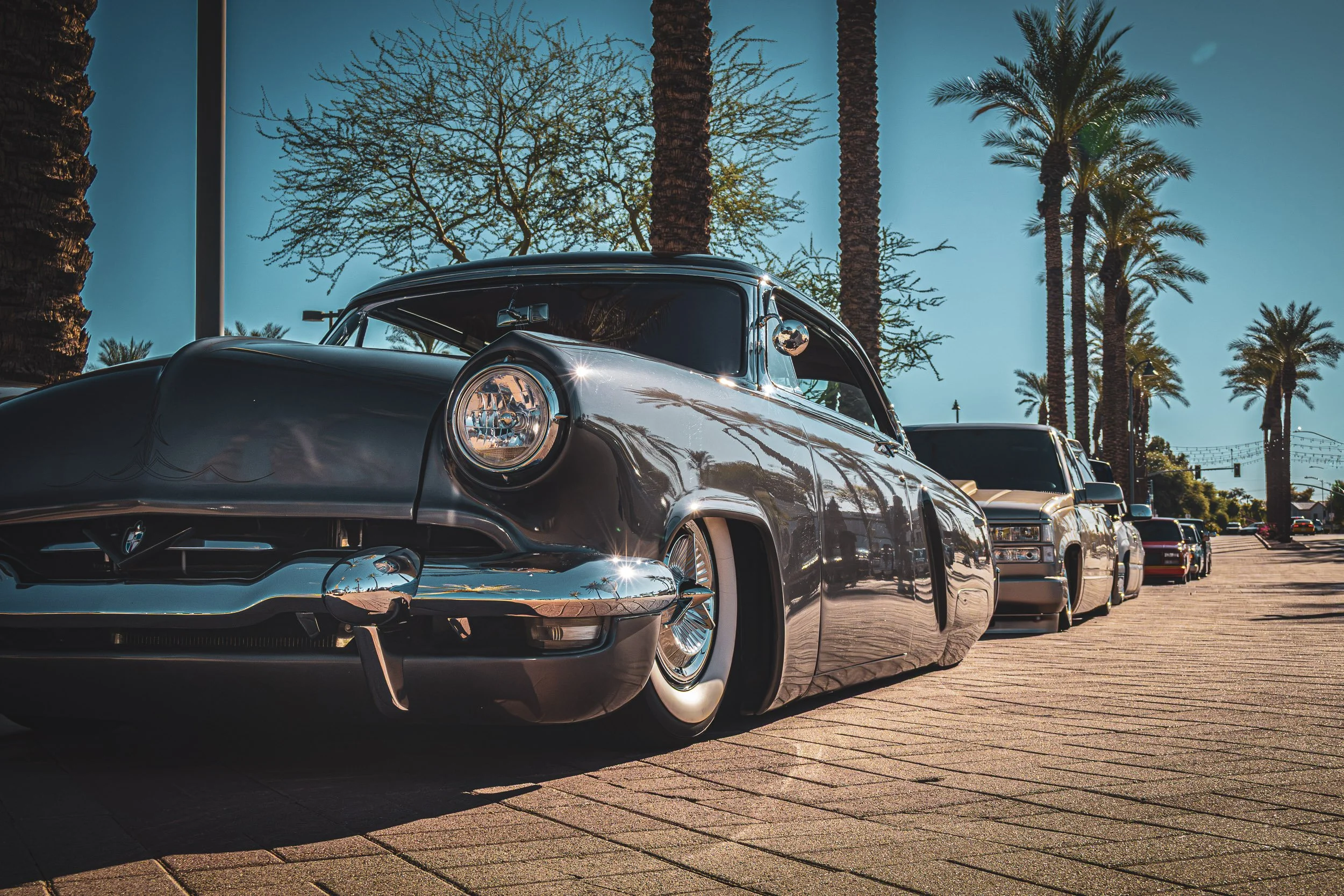 Low-angle automotive street photography highlighting the gleaming chrome bumper, whitewall tires, and wire wheels of a classic custom grey lowrider leading a lineup of modified trucks on a sunlit, palm-lined brick street in Mesa, Arizona, captured by