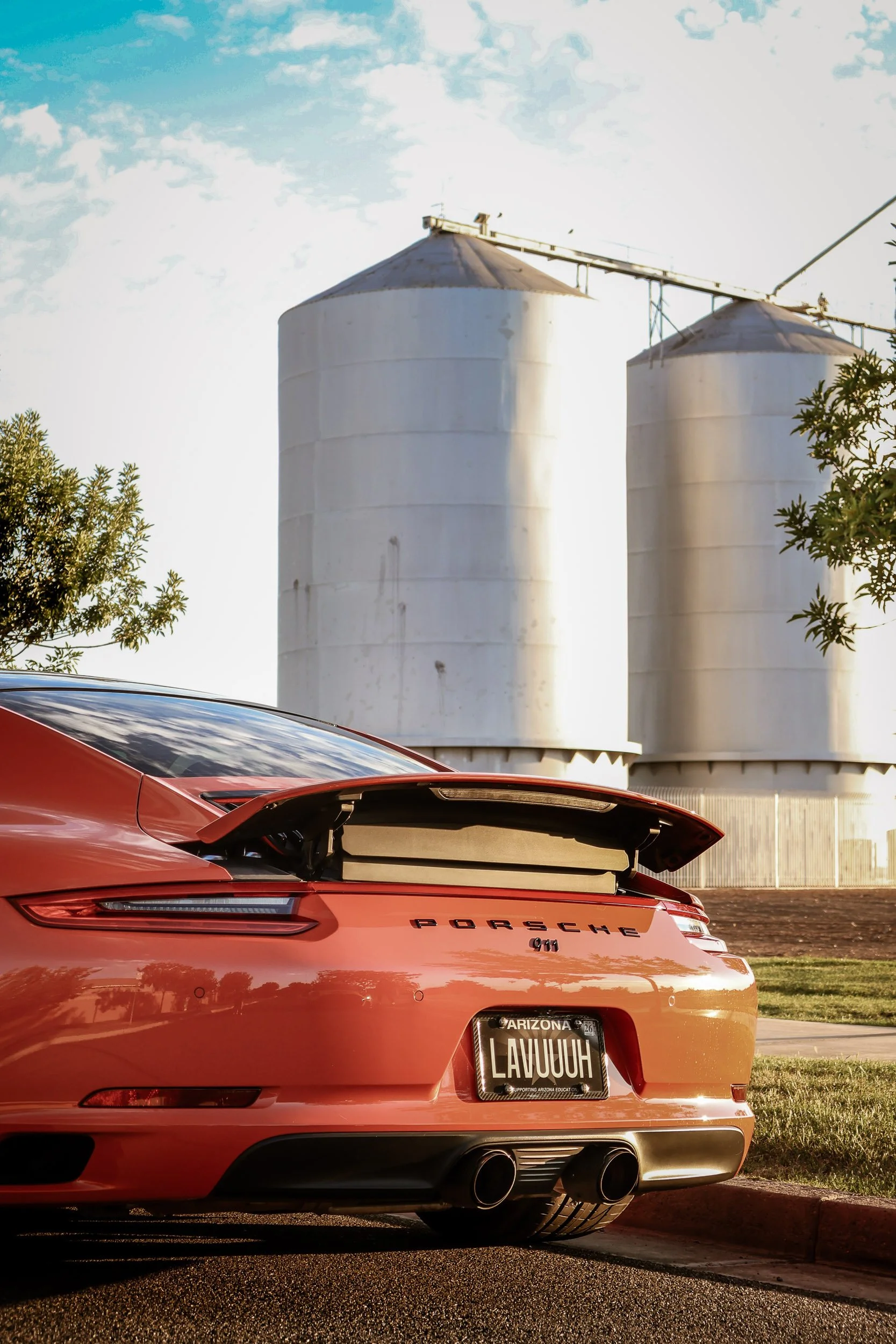 Vertical automotive photography capturing the low-angle rear profile, raised active spoiler, and center exhaust of a Lava Orange Porsche 911 C4S, framed against the historic Morrison Ranch silos in Gilbert, Arizona, captured by Curtis from Gasoline a