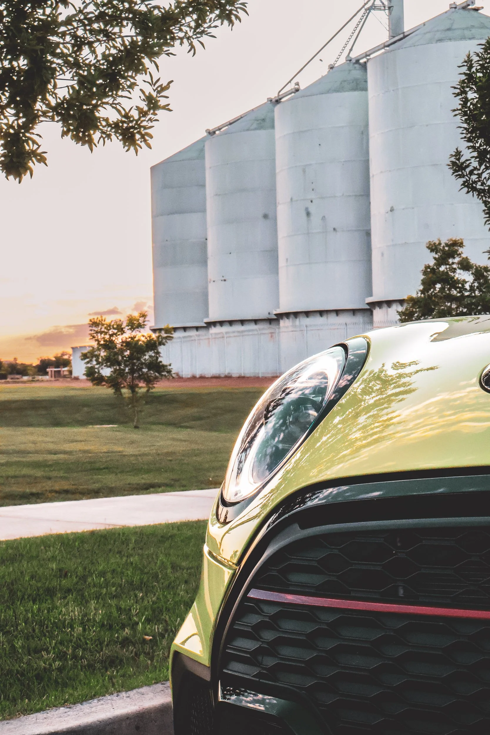 Vertical automotive photography focusing on the curved hood, reflective headlight, and honeycomb grille of a vibrant yellow Mini Cooper JCW, set against a sunset sky and the historic Morrison Ranch silos in Gilbert, Arizona, photo by Curtis at Gasoli