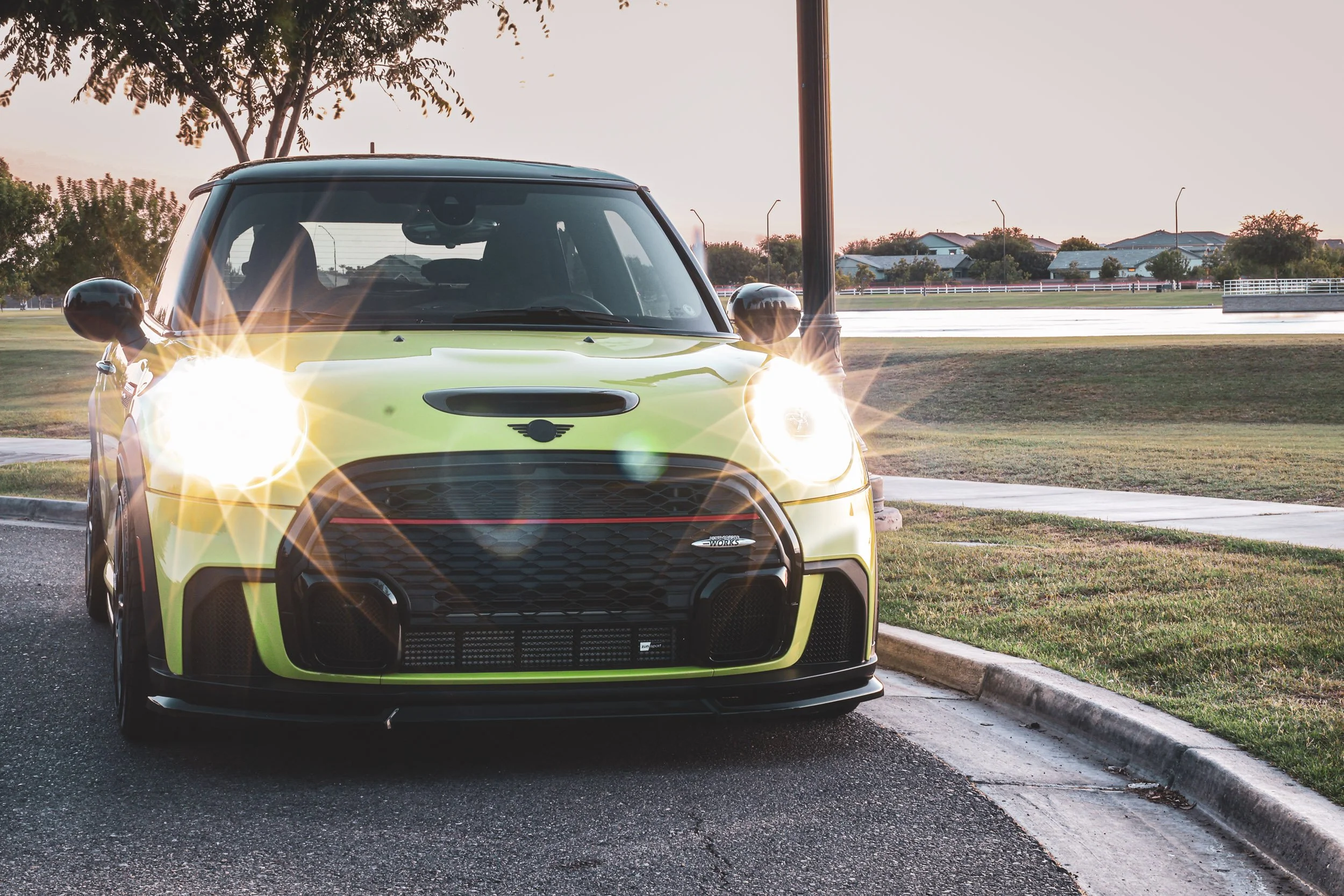 Front-facing automotive photography featuring a vibrant yellow Mini Cooper JCW with bright illuminated halo headlights, parked next to a neighborhood park near sunset in Morrison Ranch, Gilbert, Arizona, captured by Curtis from Gasoline and Dopamine.